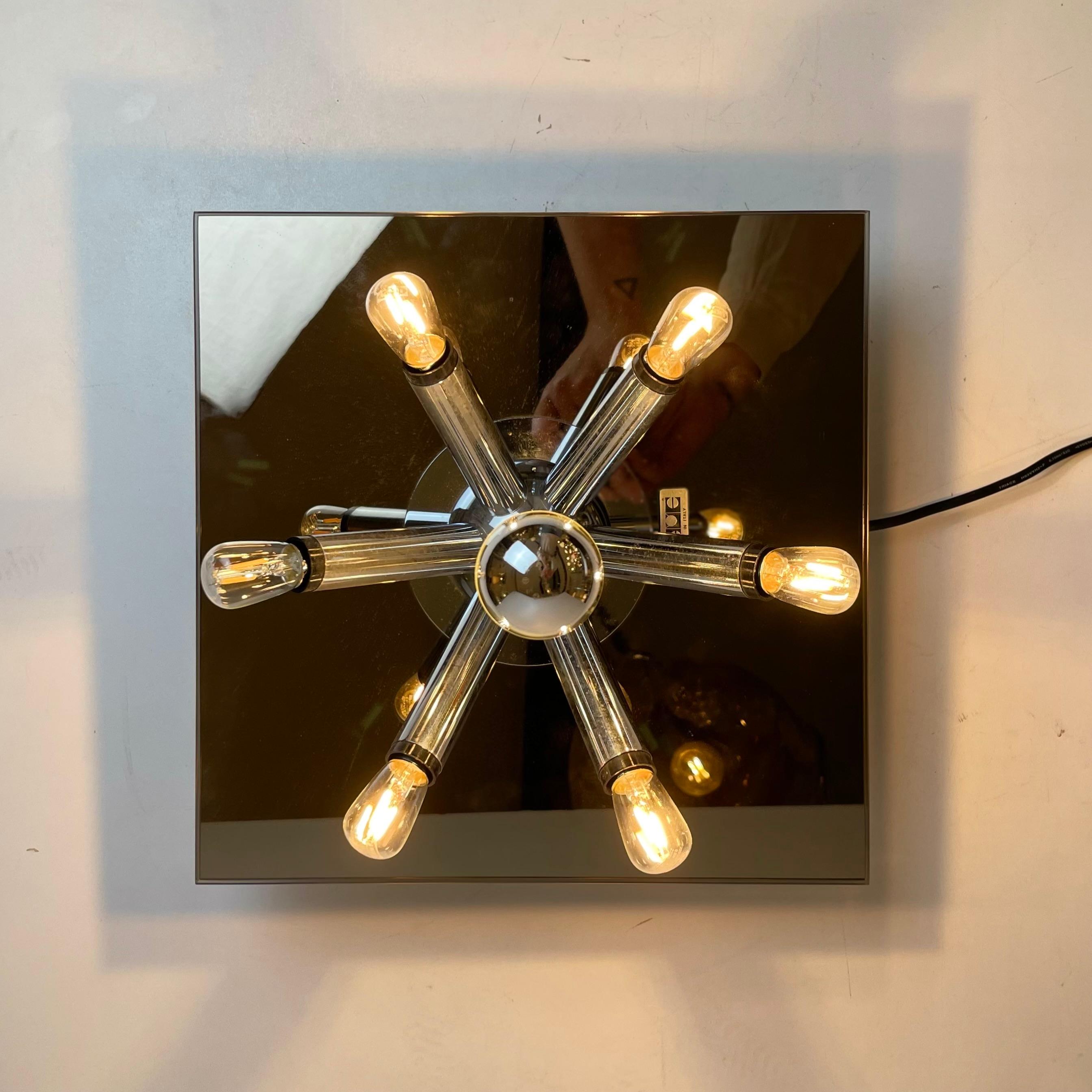 Lampada da parete e/o soffitto italiana composta da una base quadrata a specchio con sette rami in metallo cromato e sorgenti luminose. Disegnato da Bath Accessori Di Bagno negli anni '70. Viene fornito nella sua scatola originale. Vince l'Oscar