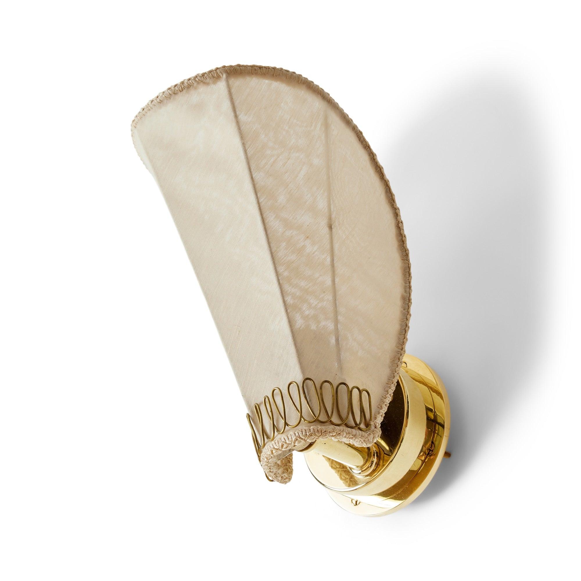 A wall mounted light fixture featuring a delicate fabric shade embellished with a gracefully twisted single piece of brass resembling flower pedals, mounted on a stepped brass disc.