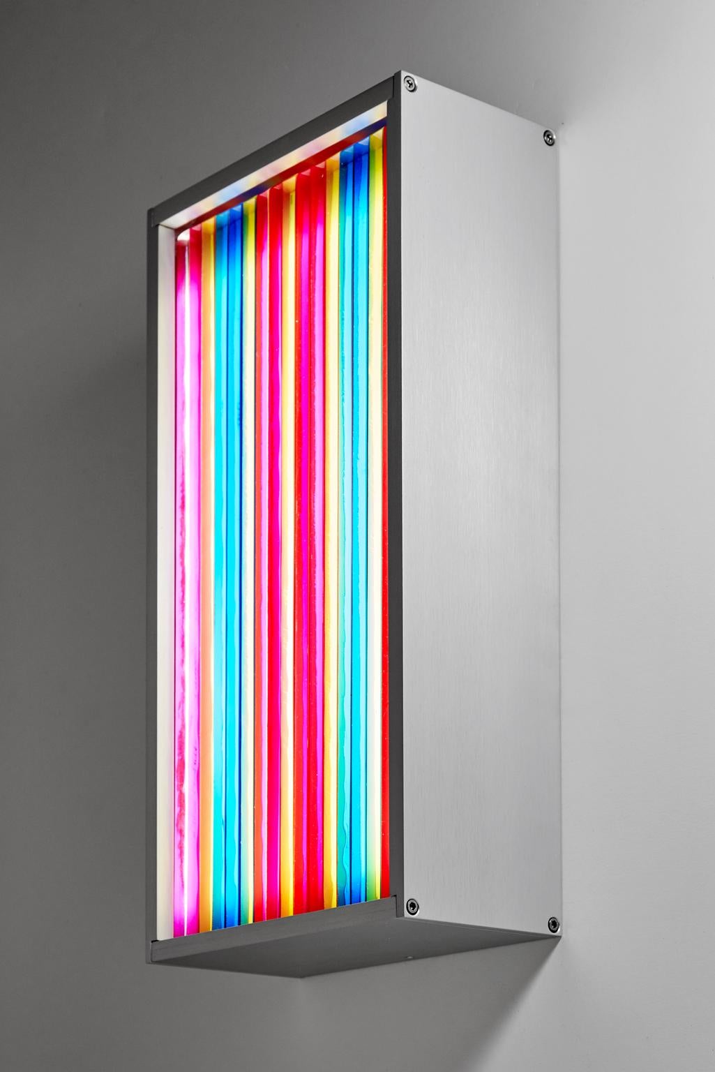 This wall sconce is created by laminating clear glass pieces together in a multicolored vertical pattern using pigmented adhesive. An LED light panel illuminates the glass and transparent pigment from behind. In a sense, the glass acts as a lens,