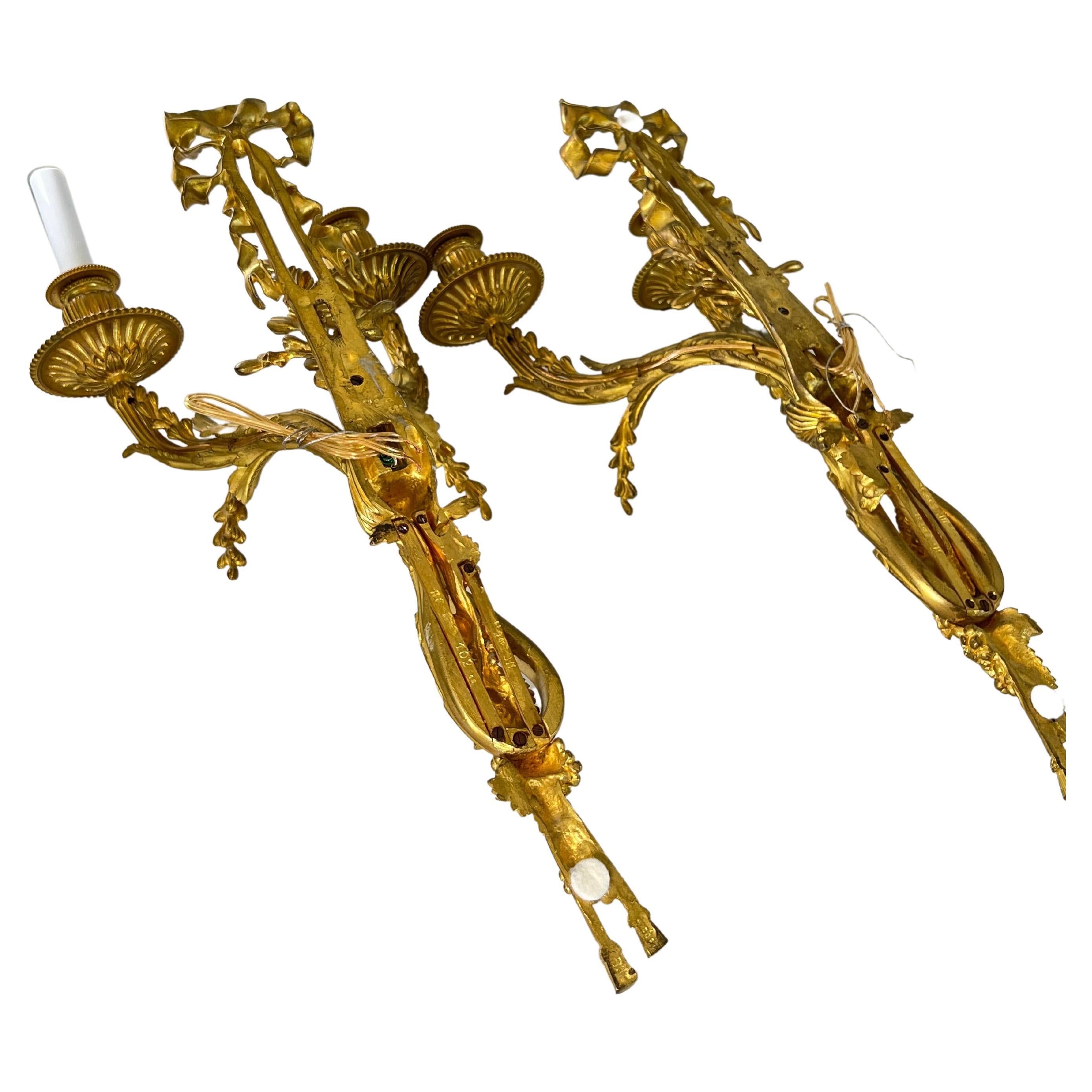 Rare pair of finely chiseled matte and shiny gilt bronze sconces. An imposing tied ribbon overhangs the sconces. A knotted link is in the center from which two berry branches come out. the bs of the sconce end with two pompoms. Along the shaft
