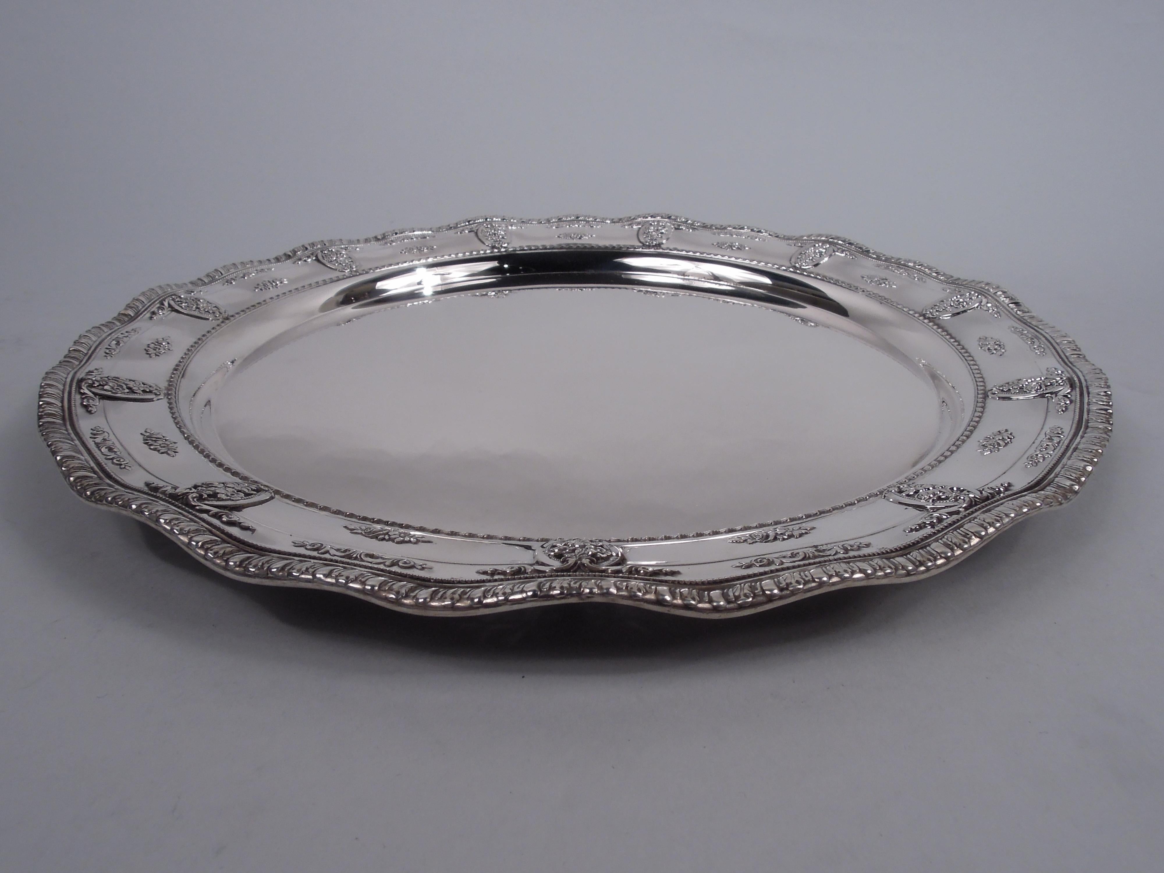 Rose Point sterling silver tray. Made by Wallace in Wallingford, Conn. Round and plain well. Tapering shoulder with open scroll ovals inset with pendant flowers alternating with chased floral clusters between beaded and gadrooned borders. Wavy