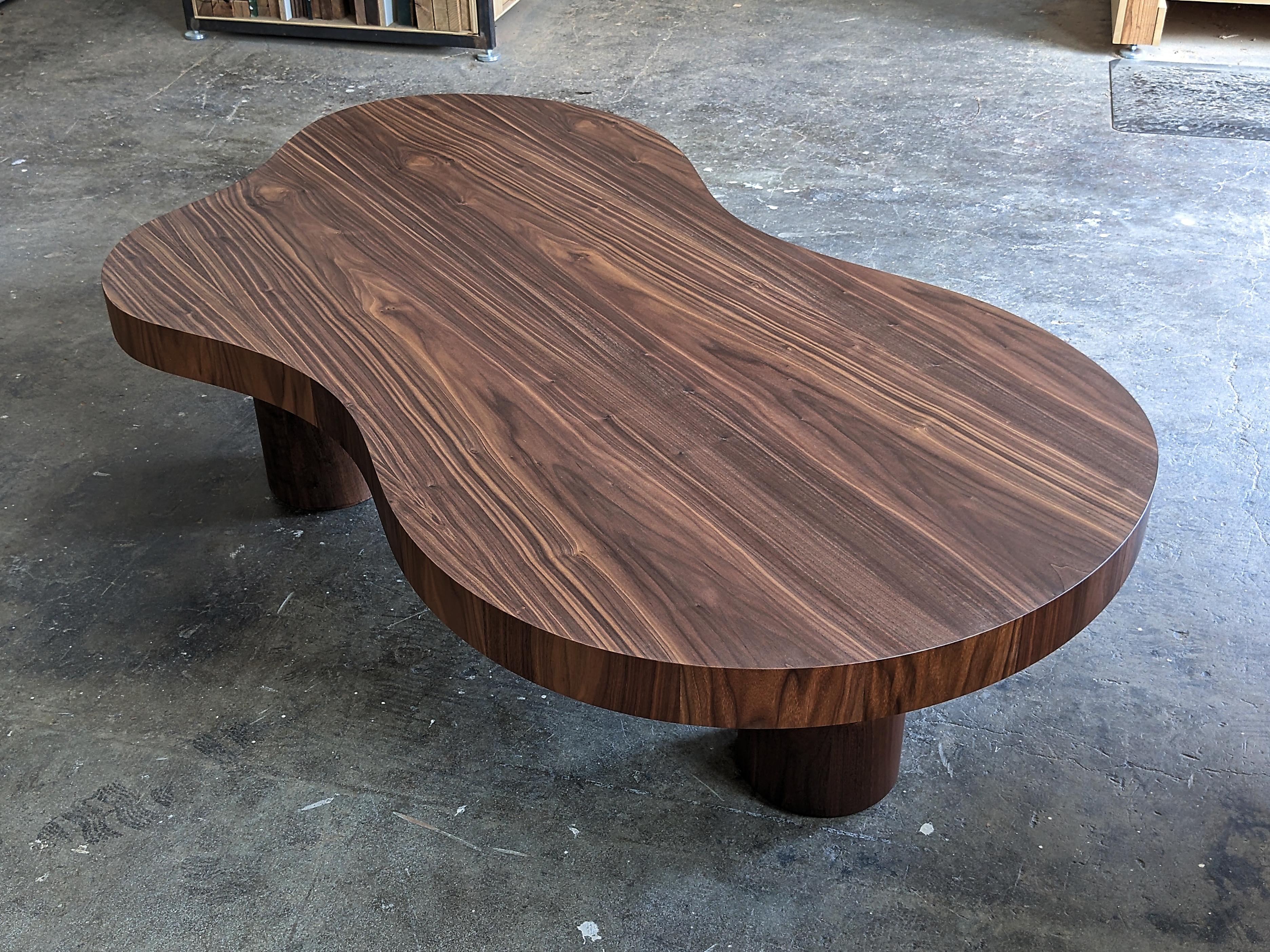 Freeform Walnut coffee table based on the style of historical pieces of the 20th century. This coffee table features an irregularly curved tabletop with vertical grain thick-veneered perimeter. Three turned solid Walnut legs support the thick