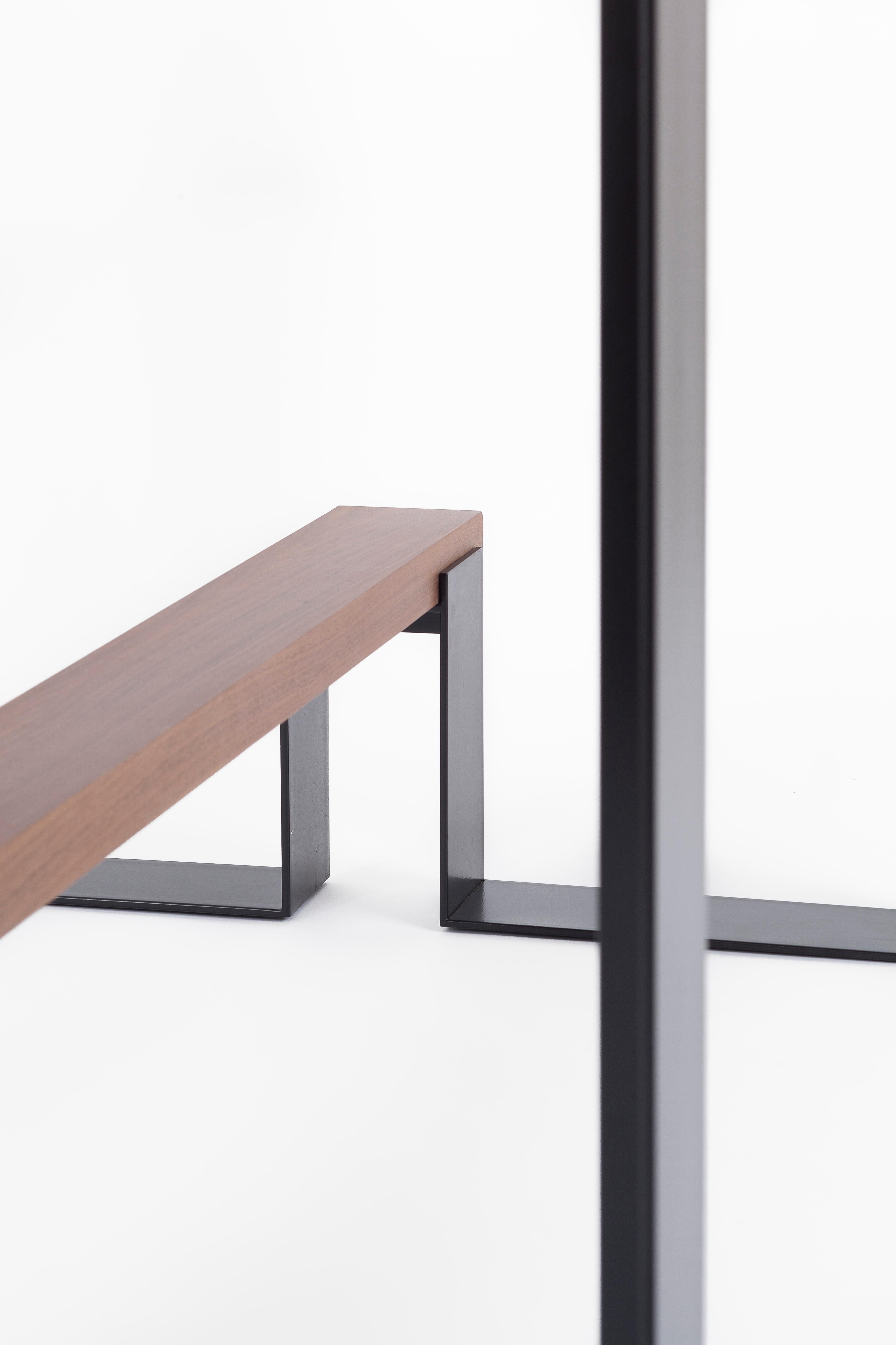 A thick (4cm) tabletop, veneered with premium wood (oak or american walnut), resting on hand-welded and polished steel brackets. An elegant footrest, forming a rich contrast with the smooth mat powder coated steel. High end materials, a slick and