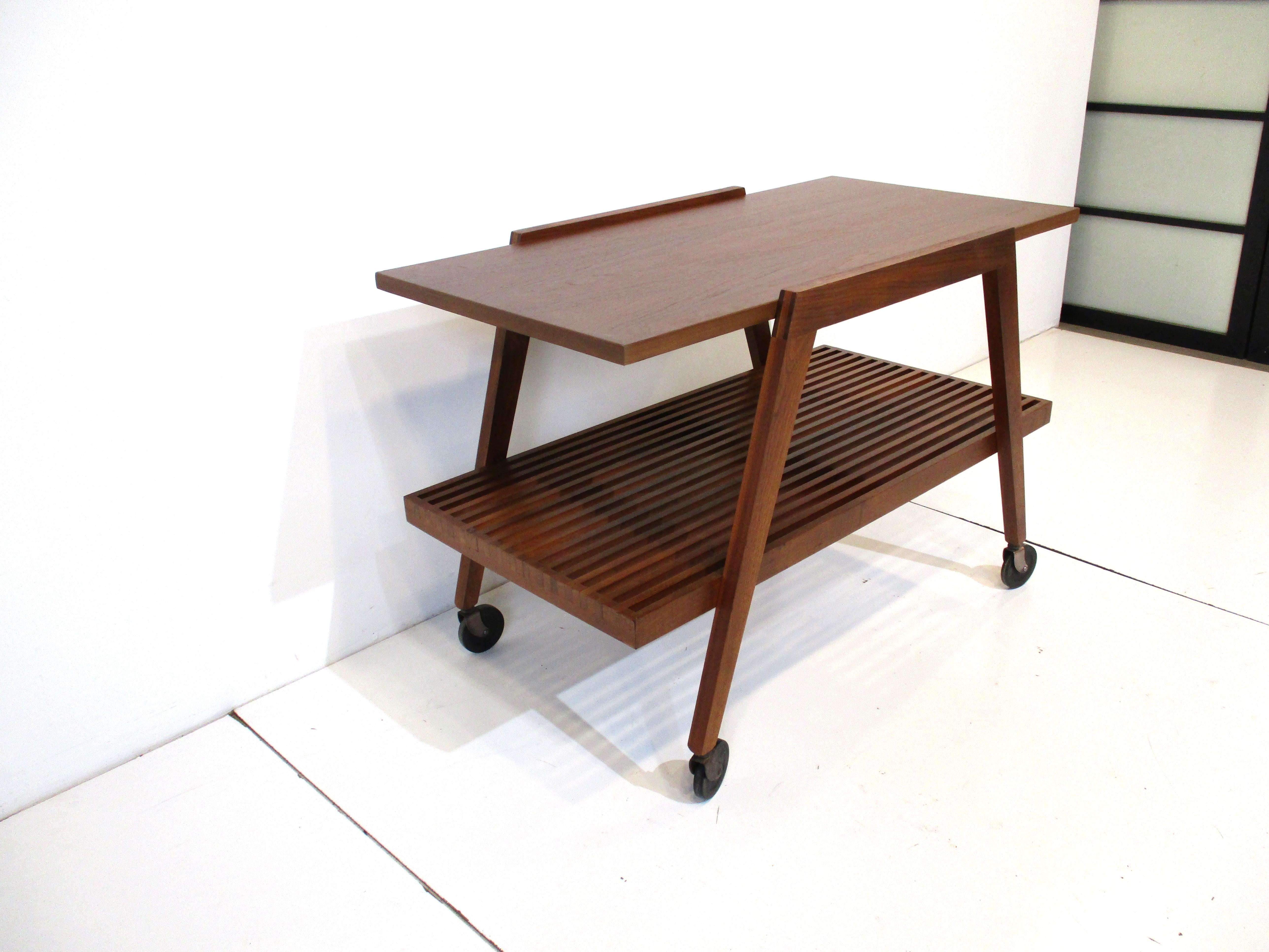 A medium dark walnut rolling bar cart with solid upper shelve and lower slat styled shelve in the manner of George Nelson. The squared arched side frames make the piece sturdy and gives the piece that mid-century well crafted quality which makes