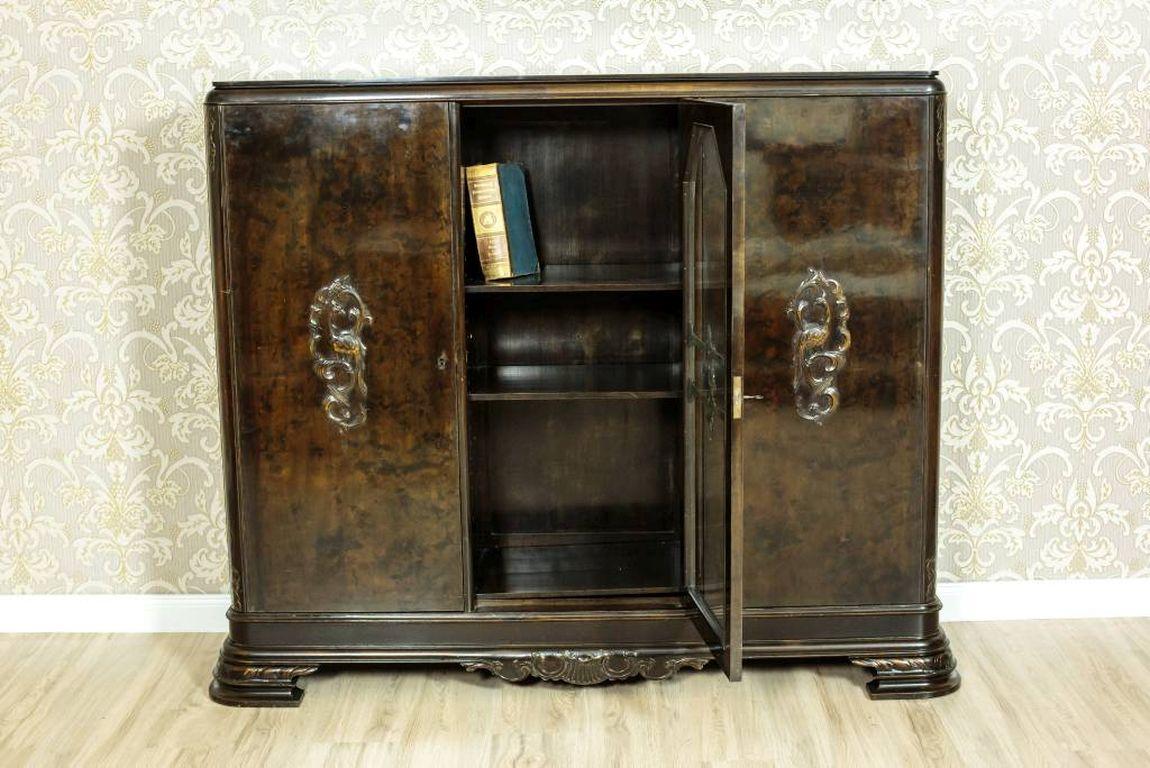 A three-door piece of furniture with a glazed central door.
The outer doors are full, with the walnut burl veneered fronts.
The legs are slightly extended beyond the plinth. The inside is divided with shelves.

Presented bookcase needs to be