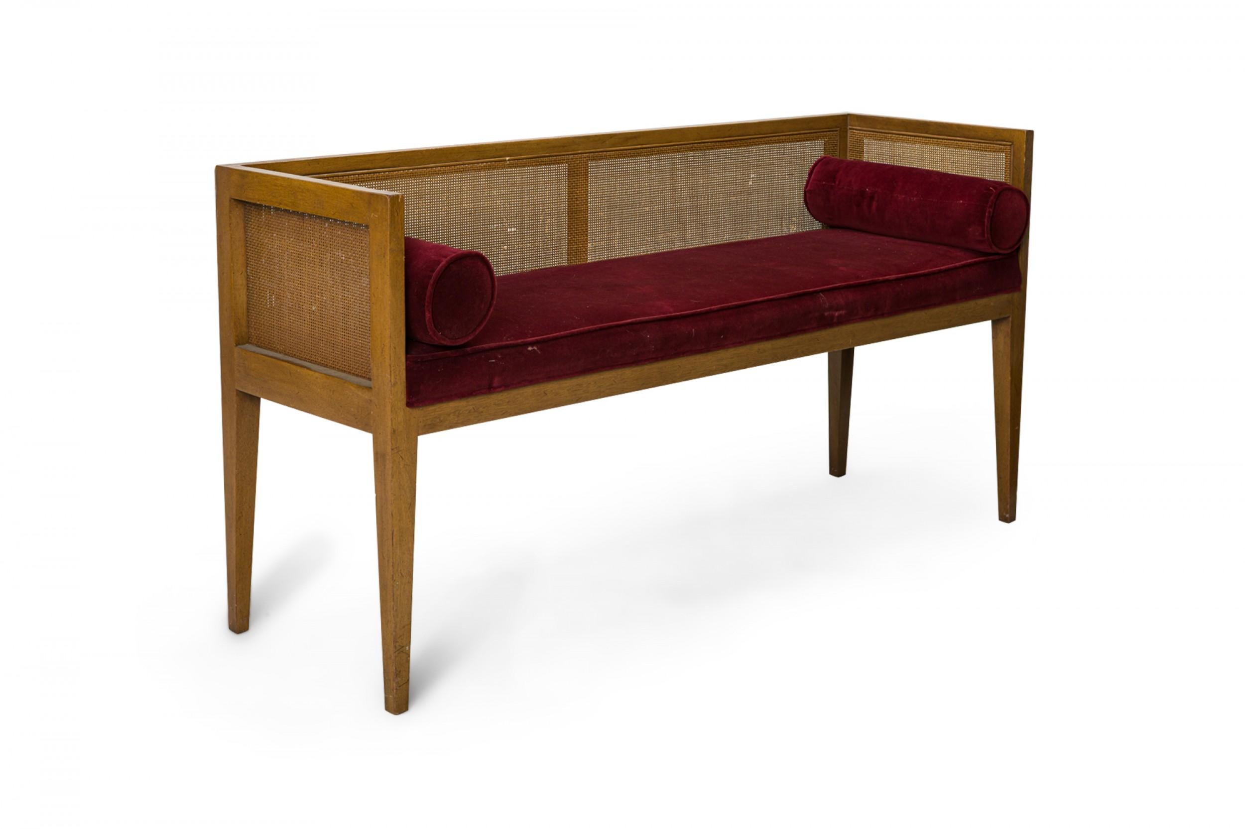 American mid-century entry bench with a rectangular walnut frame with caned back and sides, with an upholstered seat cushion in a deep burgundy velour upholstery with matching piping and two matching bolster pillows.