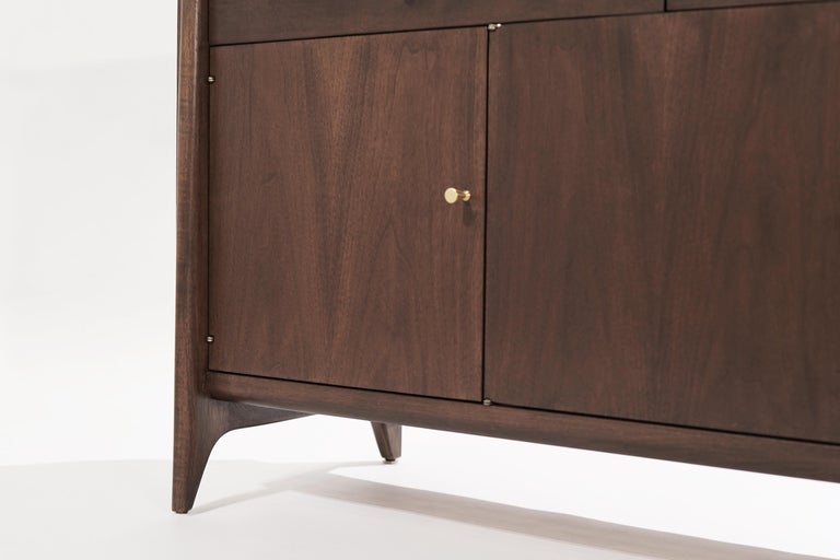 A console table or sideboard in walnut by John Stuart. Executed in walnut, featuring slopping curves and sudden sharpness. The two top drawers can be used for silverware storage or keys, the bottom doors open up to reveal shelving. Completely