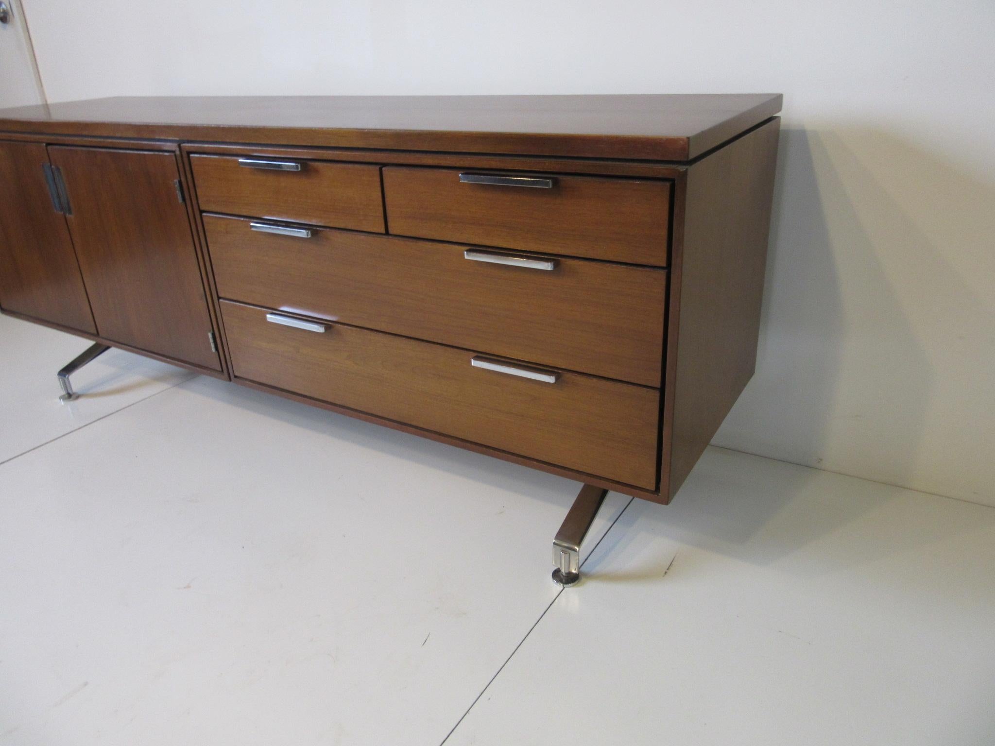 A very well constructed walnut credenza with chrome split legs and round foot pads complemented by long matching chrome pulls to the four drawers. The other side has two doors and a single shelve. The backside is finished in the same walnut so it