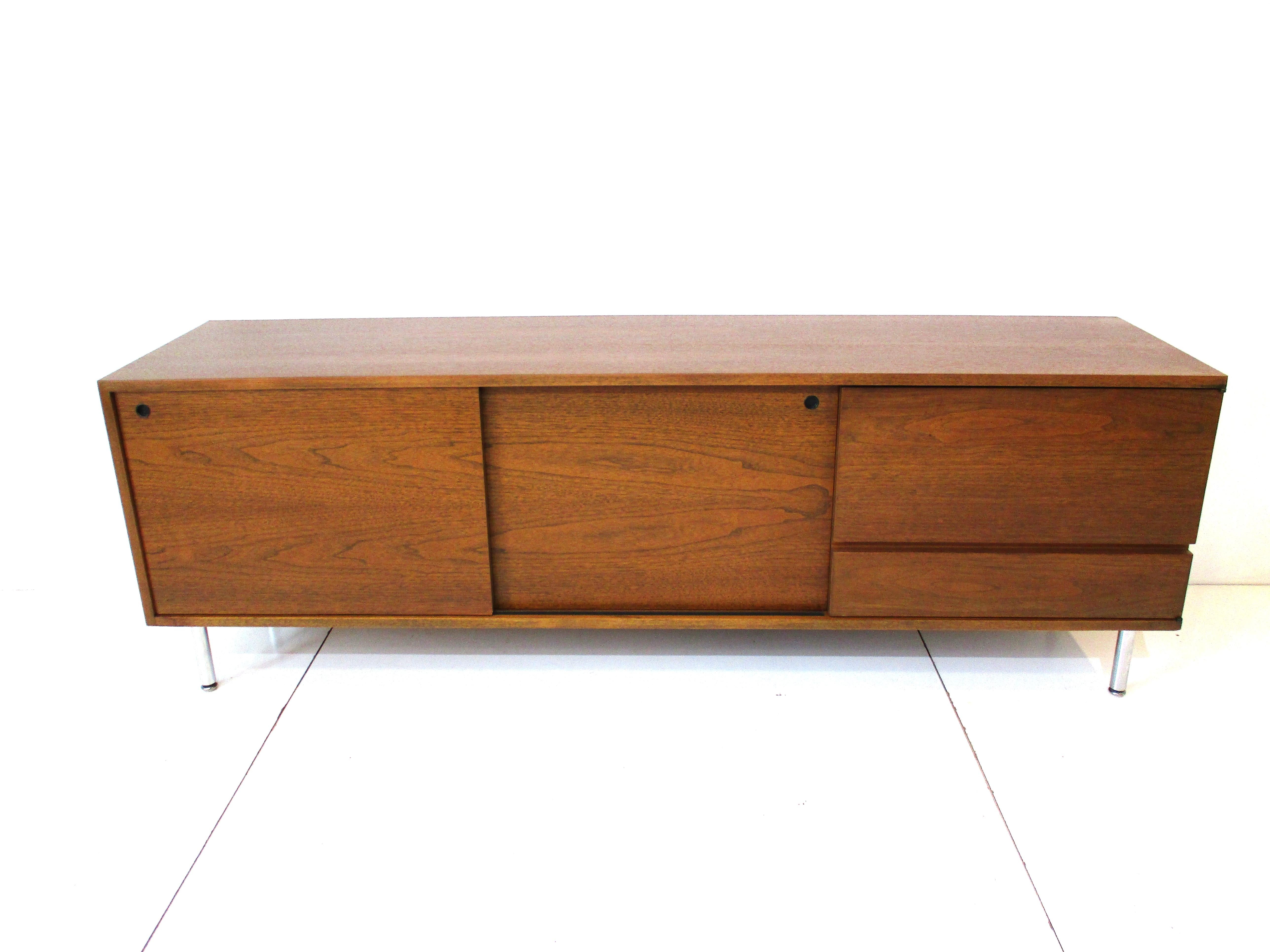 A mid century walnut credenza and two sliding doors having finger pulls in stain black with adjustable shelves inside. The one end has a door with cut in lower handle revealing a smaller drawer inside and a small adjustable shelve with lower