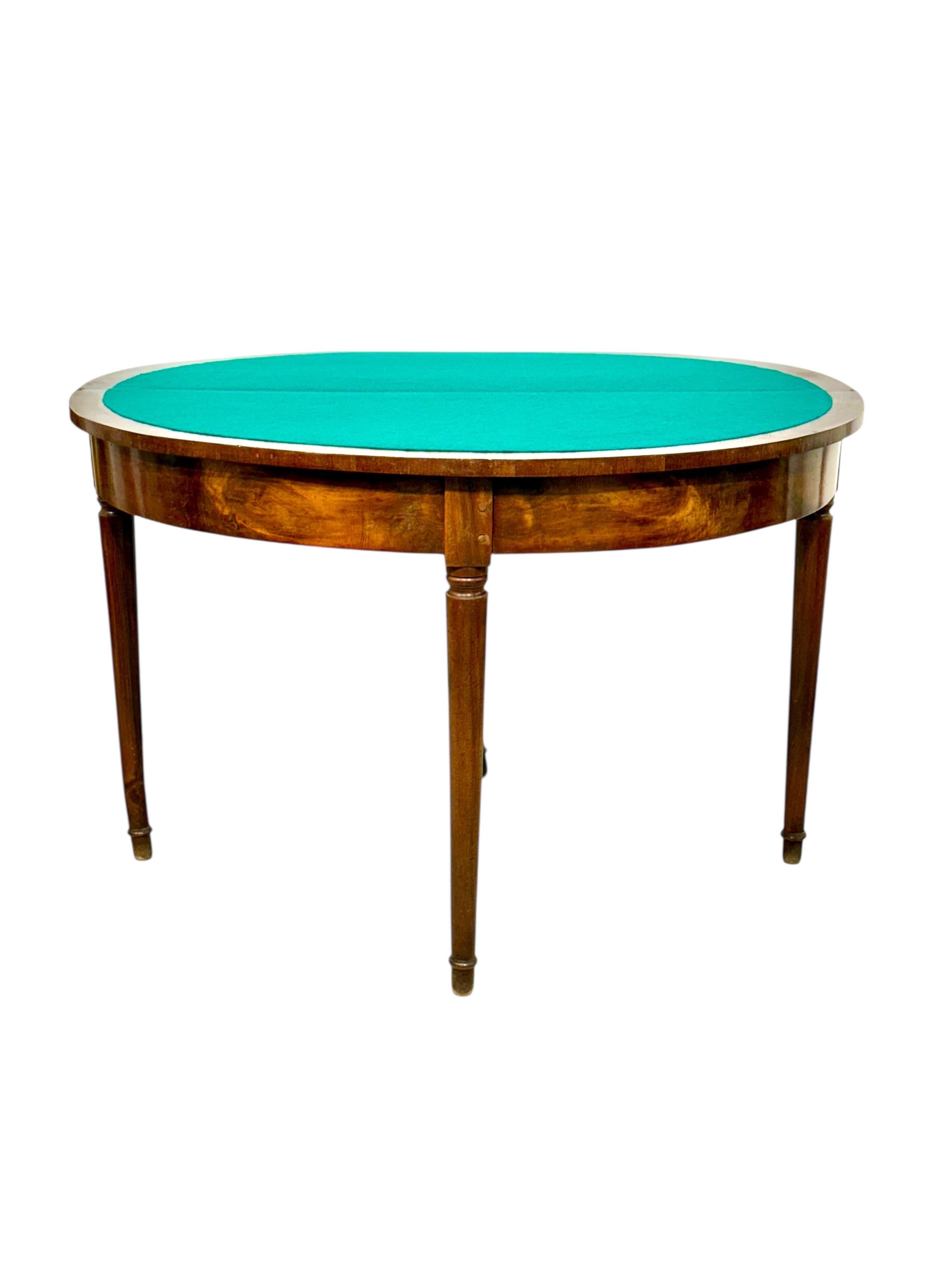 Esta elegante mesa de juego demiluna combina encanto y funcionalidad, mostrando la artesanía de los muebles de finales del siglo XVIII. Fabricada en madera maciza y chapa de nogal, la mesa descansa sobre cuatro patas esbeltas y ahusadas con sutiles