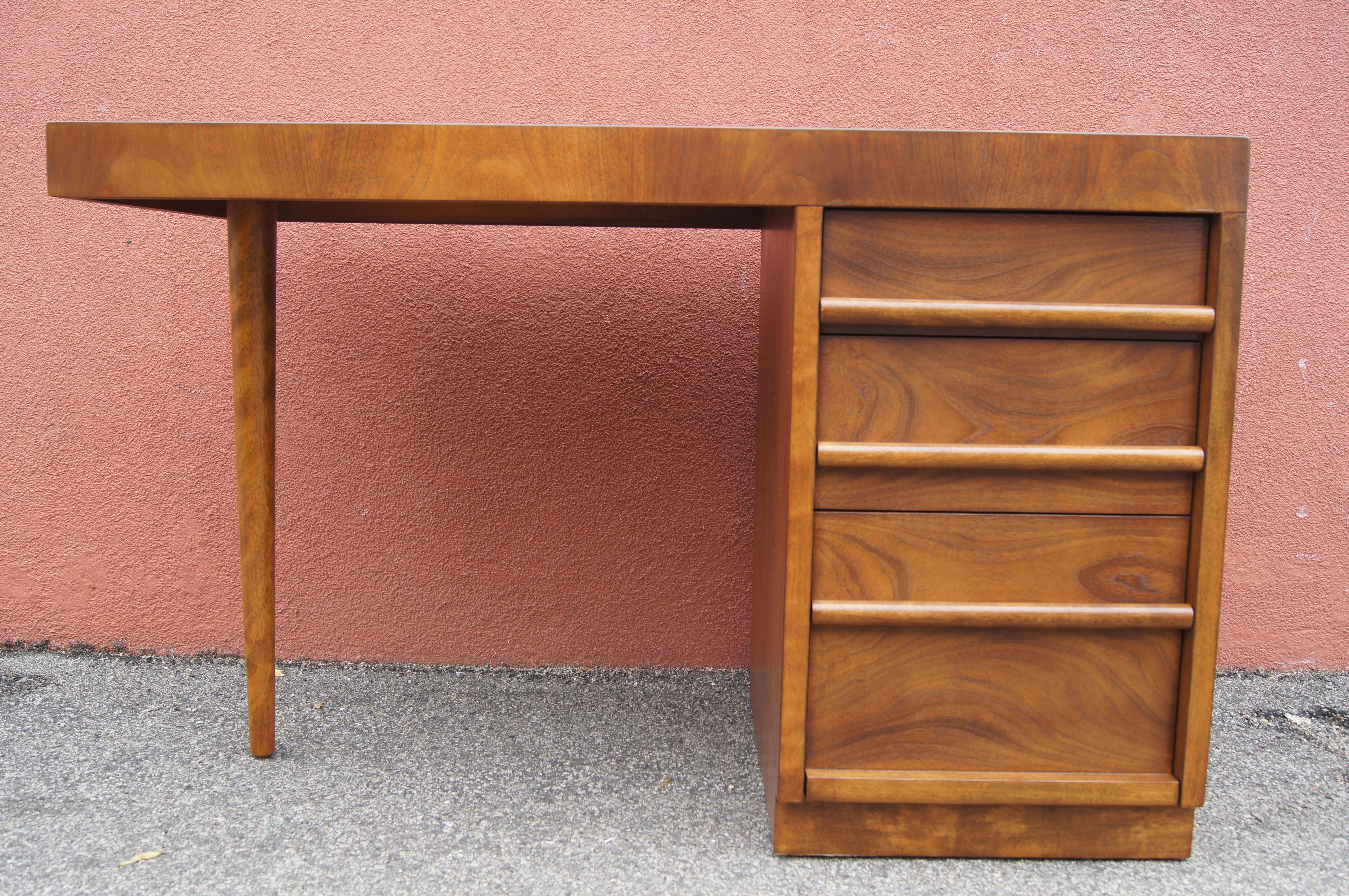 T.H. Robsjohn-Gibbings designed this walnut desk for Widdicomb. A stack of three drawers is balanced by a thick top and a single tapering leg. The divided drawers, the lowest of which is file deep, feature solid rounded pulls. The back of the desk