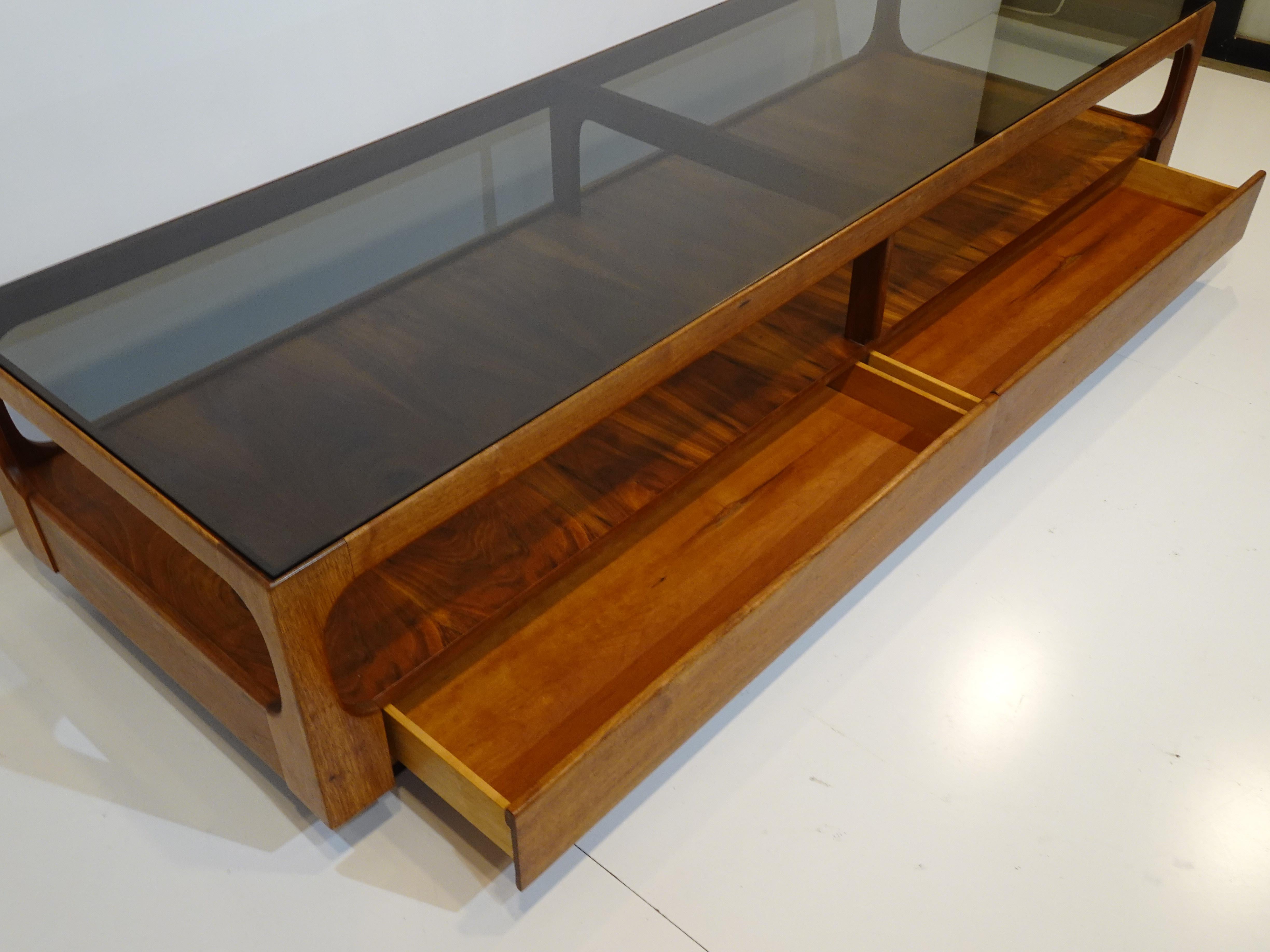 A well crafted walnut coffee table in a long cube style having well grained dark walnut to the lower shelve area . Topped with smoked glass, curved walnut edges the piece has a floating feel on it's satin black kick base and to the front are two