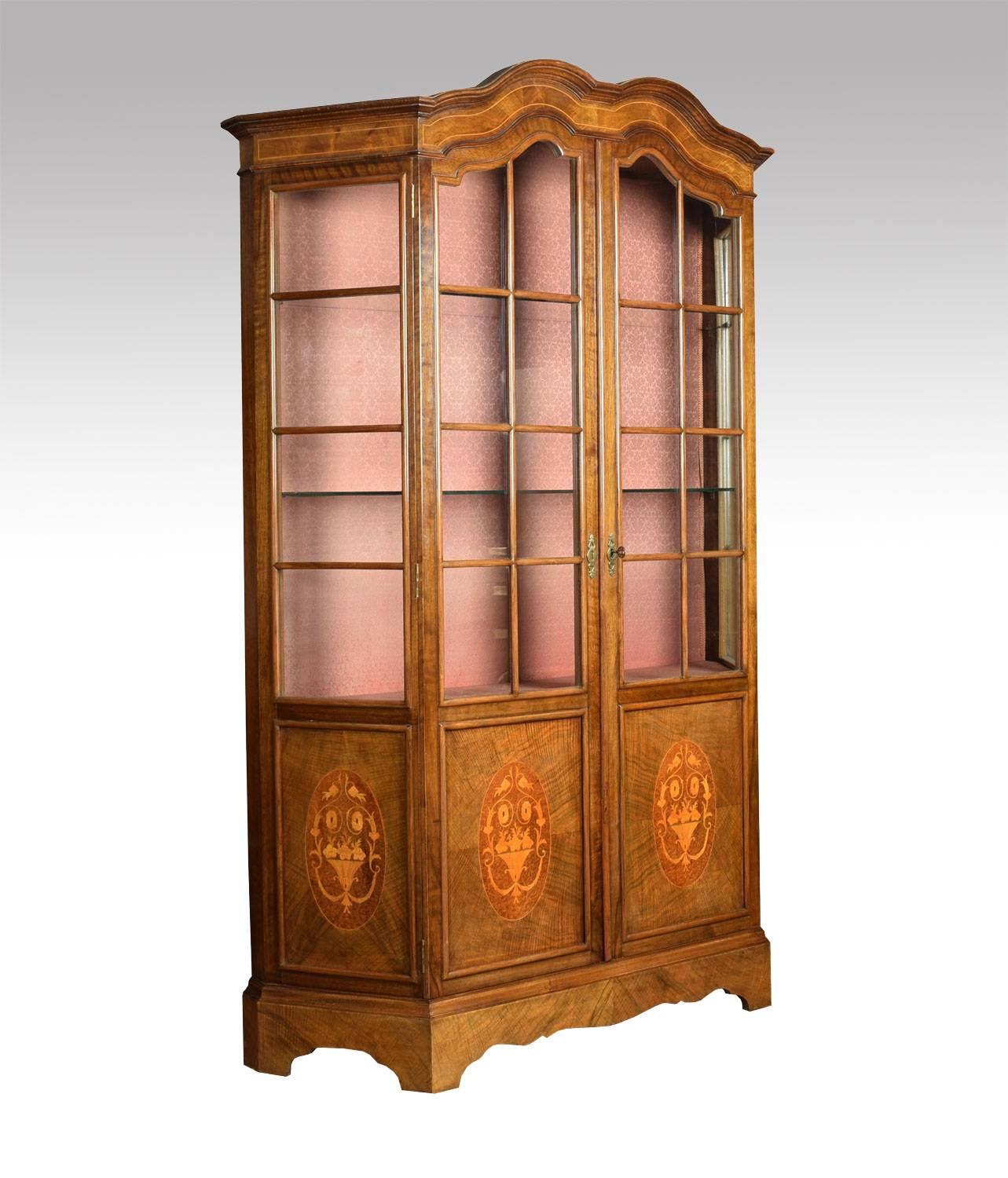 Walnut display cabinet the arched top over two central doors opening to reveal serviceable upholstered interior with two glazed shelves. Flanked by unusual concaved glass panelled sides the base section with oval inlaid panels all raised up shaped