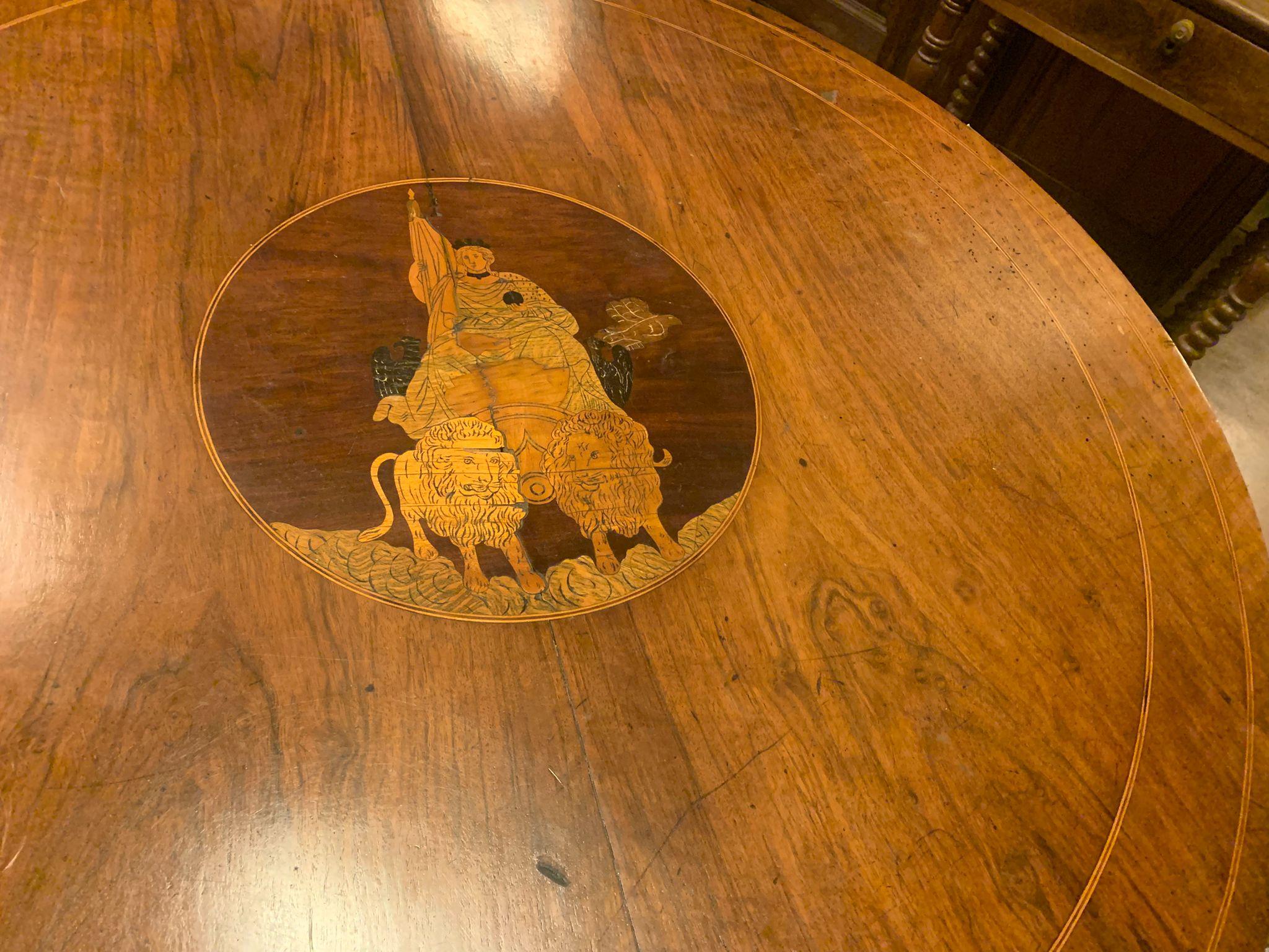 Mesa antigua de salón o comedor, de madera maciza de nogal, con tablero redondo con marquetería central, bloque central con tres pies terminados en rizo. Adornado con incrustaciones laterales donde hay 4 cajones pequeños. La incrustación de la parte