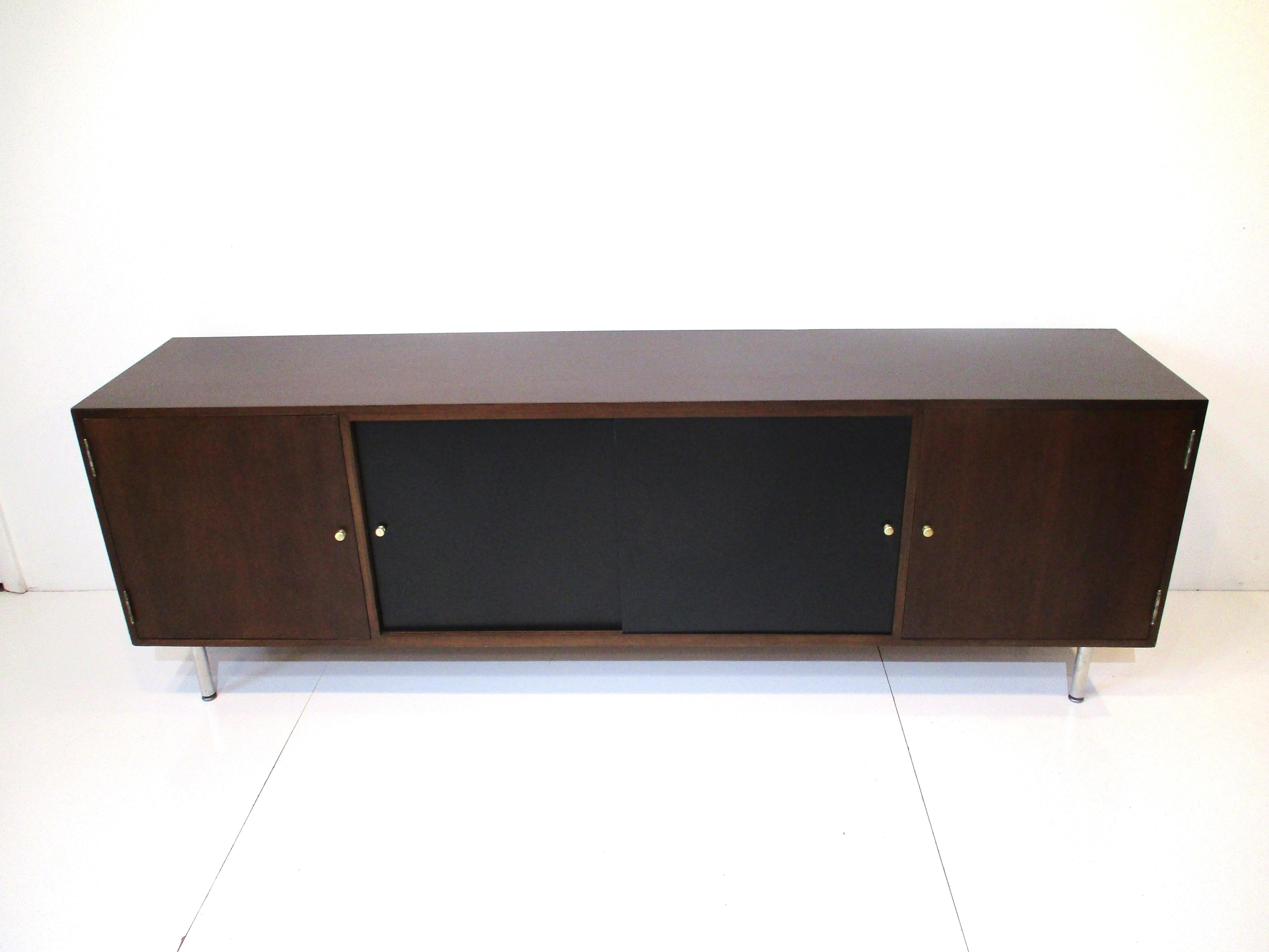 A Mid Century walnut credenza with a door to each side and two satin black sliding doors to the center all with brass pulls . Great storage with fixed shelves to each section and sitting on nickel toned metal legs in the manner of George Nelson and