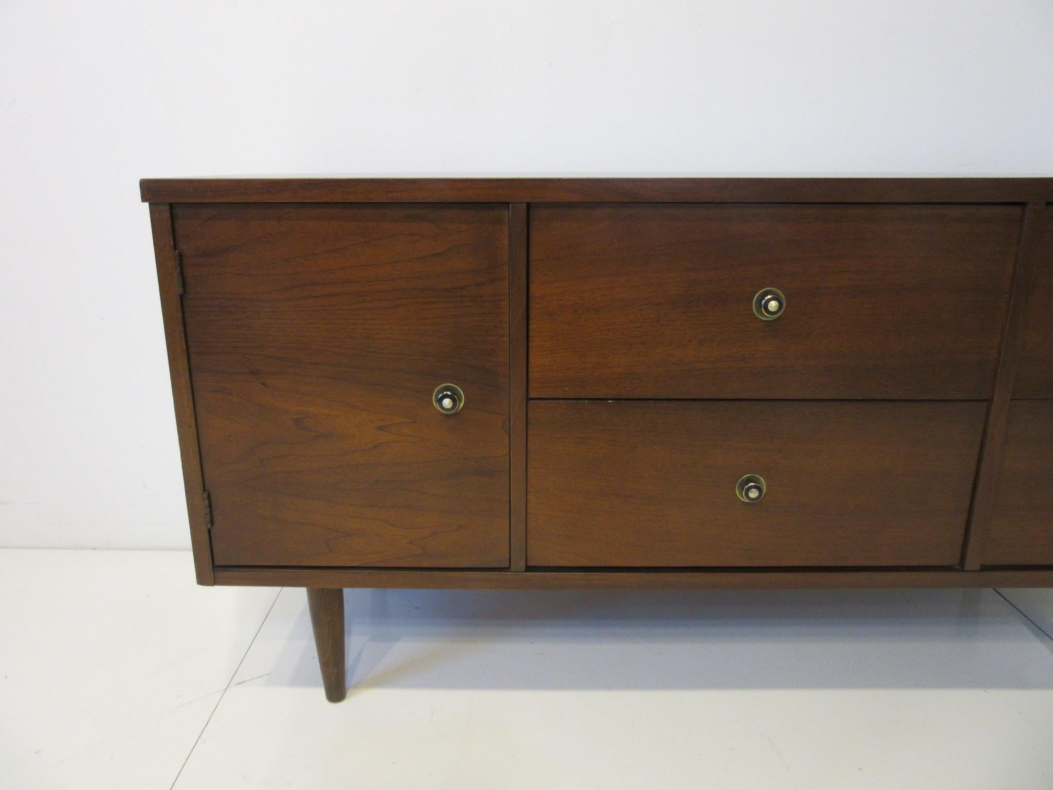 A smaller sized credenza / sever / cabinet with door to the left having a shelve and four drawers one with dividers. Black and brass pulls to the front and sitting on conical legs, retains the original manufactures label by John A. Schwarz,