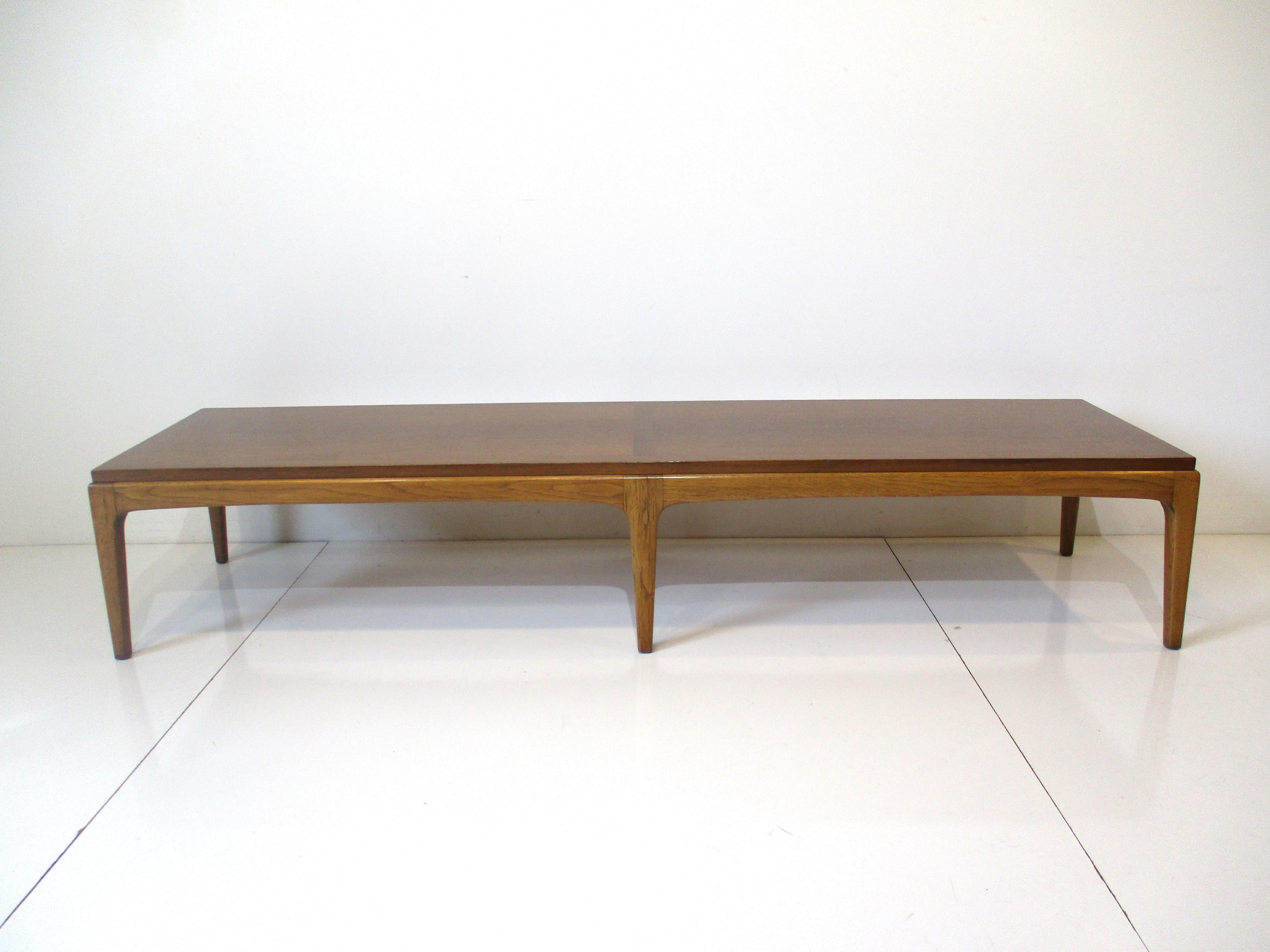 A well grained walnut topped long coffee table with lower legs and details in a medium mahogany. Slim tapered legs give the table a lighter feel from the Rhythm Collection by Altivista Lane.