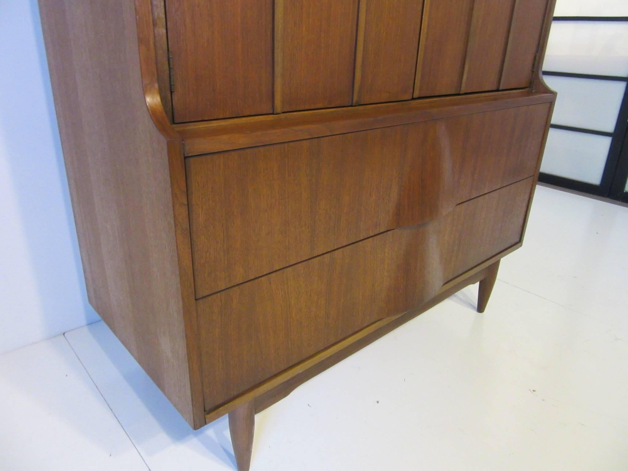 A walnut midcentury chest with two upper doors opening to reveal three drawers and lower two drawers with sculptural fronts that have built in pulls and plenty of storage . A very attractive design with conical legs and lower stretchers, in the