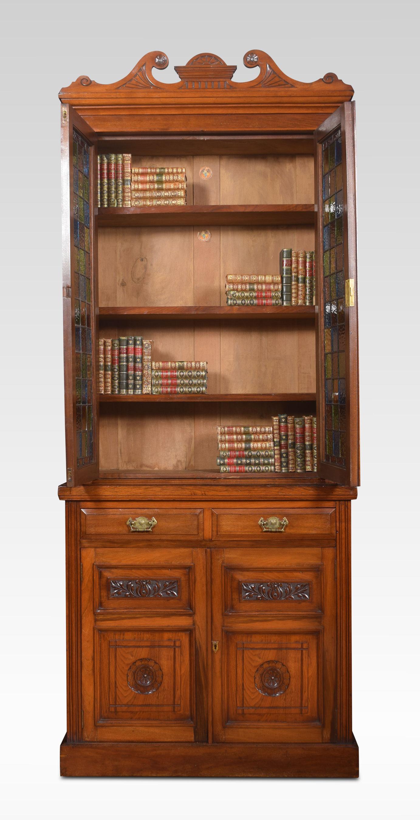 Walnut narrow bookcase of narrow proportions. The carved cornice above a pair of leaded stained glass door enclosing adjustable shelved interior. Flanked by reeded columns. To the base with two frieze drawer and a panelled cupboard doors below