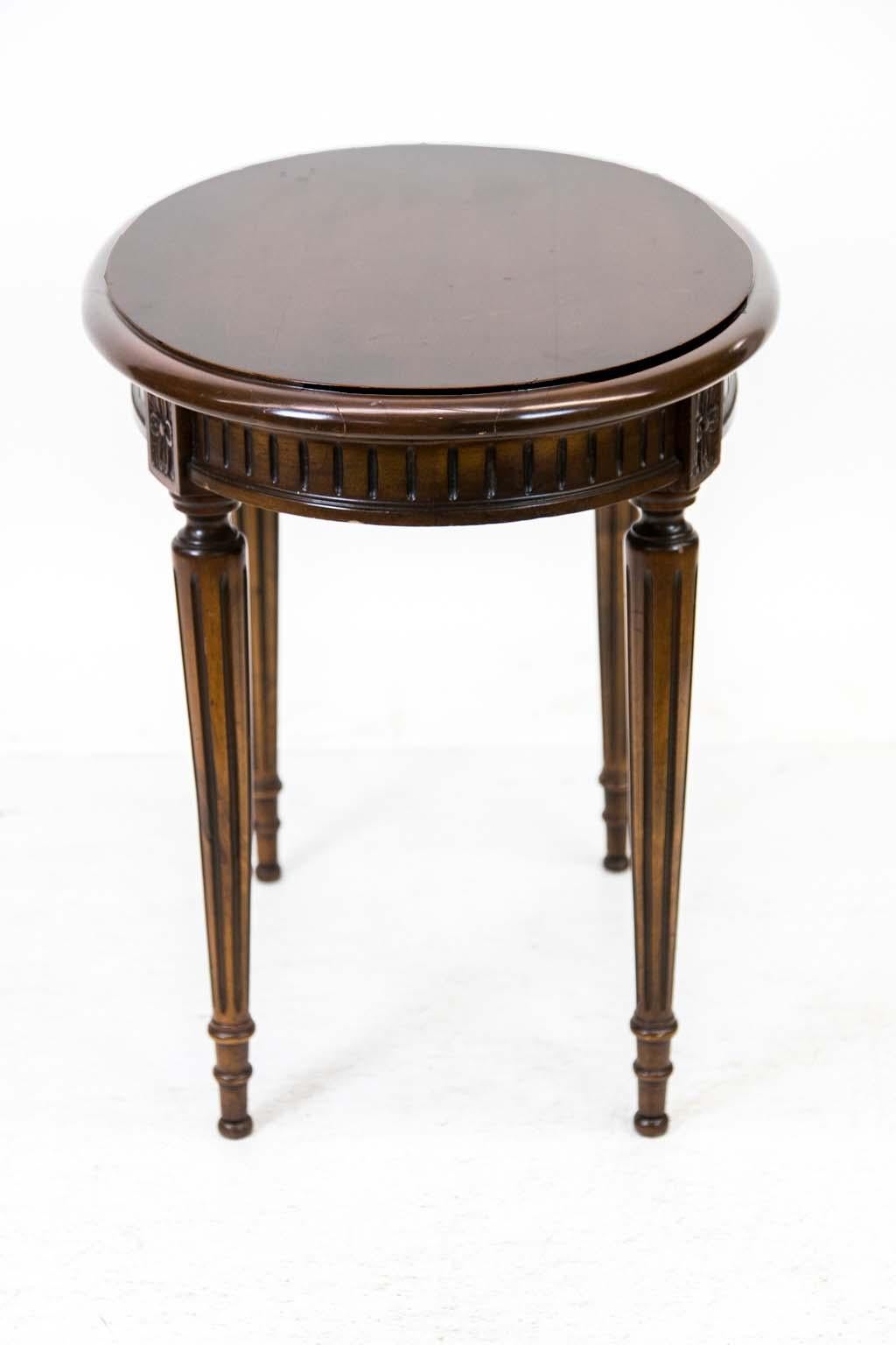 This walnut oval center table has a bull nosed edge molding with a fluted apron and legs that are also fluted. There is a carved floral panel at the top of the apron. The top finish has noticeable crazing.
  
