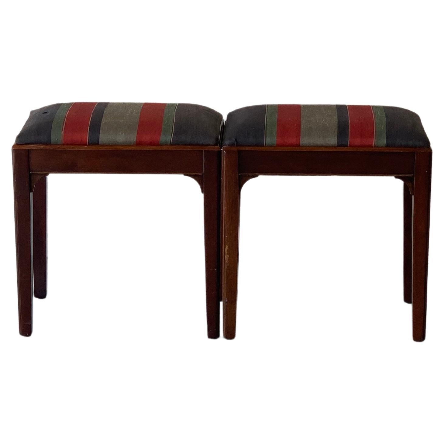 A pair of 1960' s walnut bench stools in original vintage roman stripe upholstery. Wood frame is in excellent condition. Fabric on one stool has a minor upholstery tear. Colors are a variation of subdued blues, reds & green.
