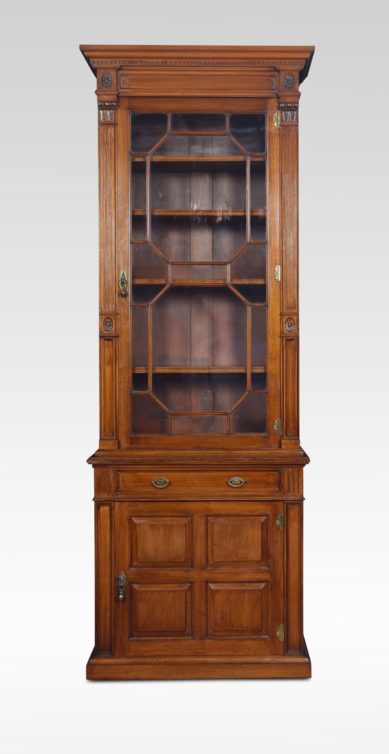 Walnut pier bookcase of narrow proportions, in the manner of Howard & Sons. The dentil cornice above an astragal glazed door enclosing adjustable shelved interior. Flanked by reeded pilasters with lotus capitals. To the base with a frieze drawer and