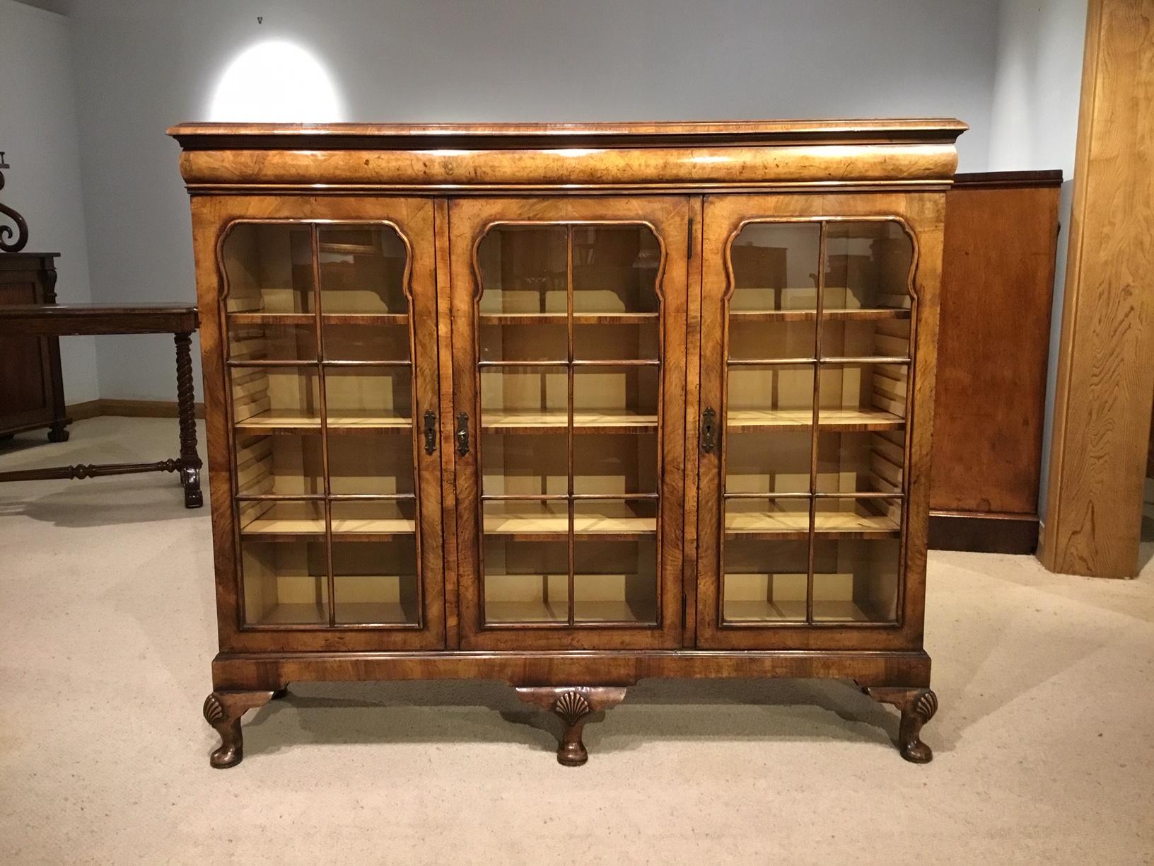 A walnut Queen Anne Revival three-door bookcase. The rectangular top veneered in beautifully figured burr walnut with block moulding and a cushion shaped frieze. The front with three walnut veneered glazed doors opening to reveal nine adjustable