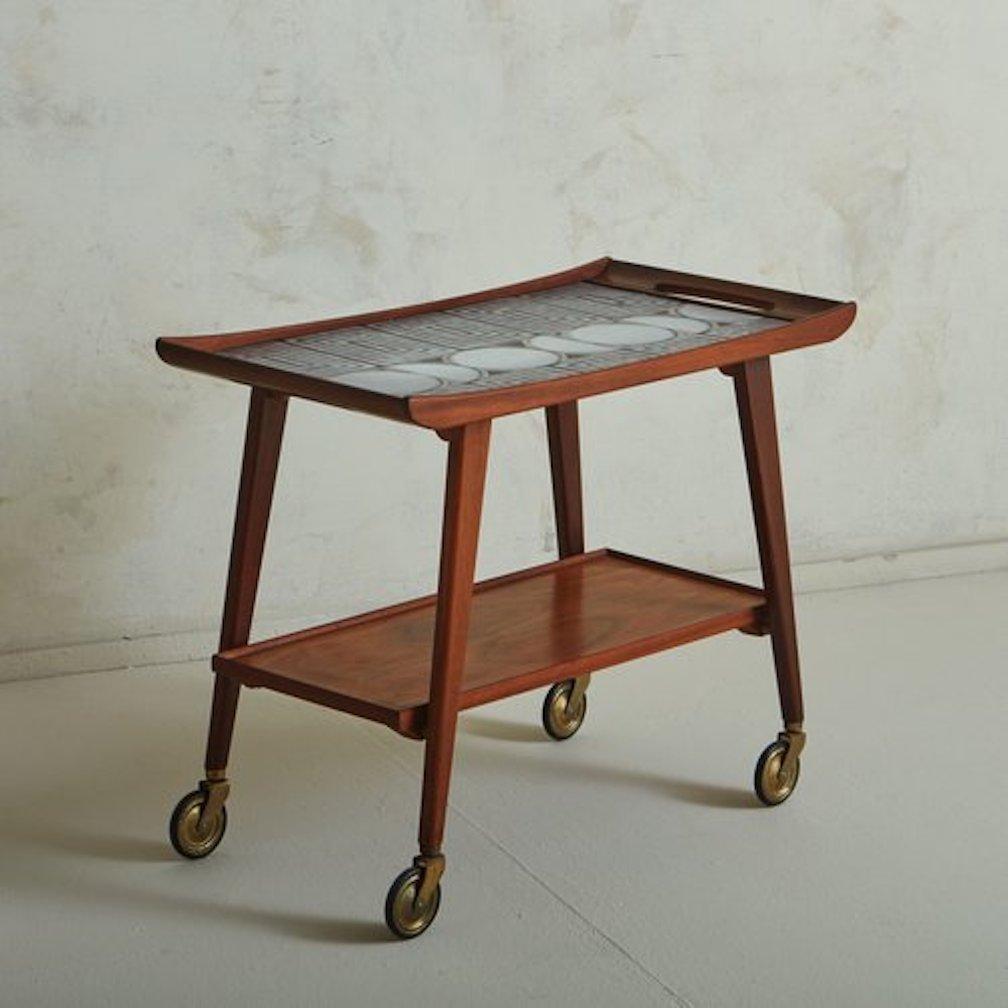 A 1960s German serving trolley by Opal Kleinmöbel. This piece features a handsome two-tier walnut veneer frame with elegantly curved sides and cut out handles. It has an inlaid ceramic tile tabletop with a geometric pattern in blue, black and white