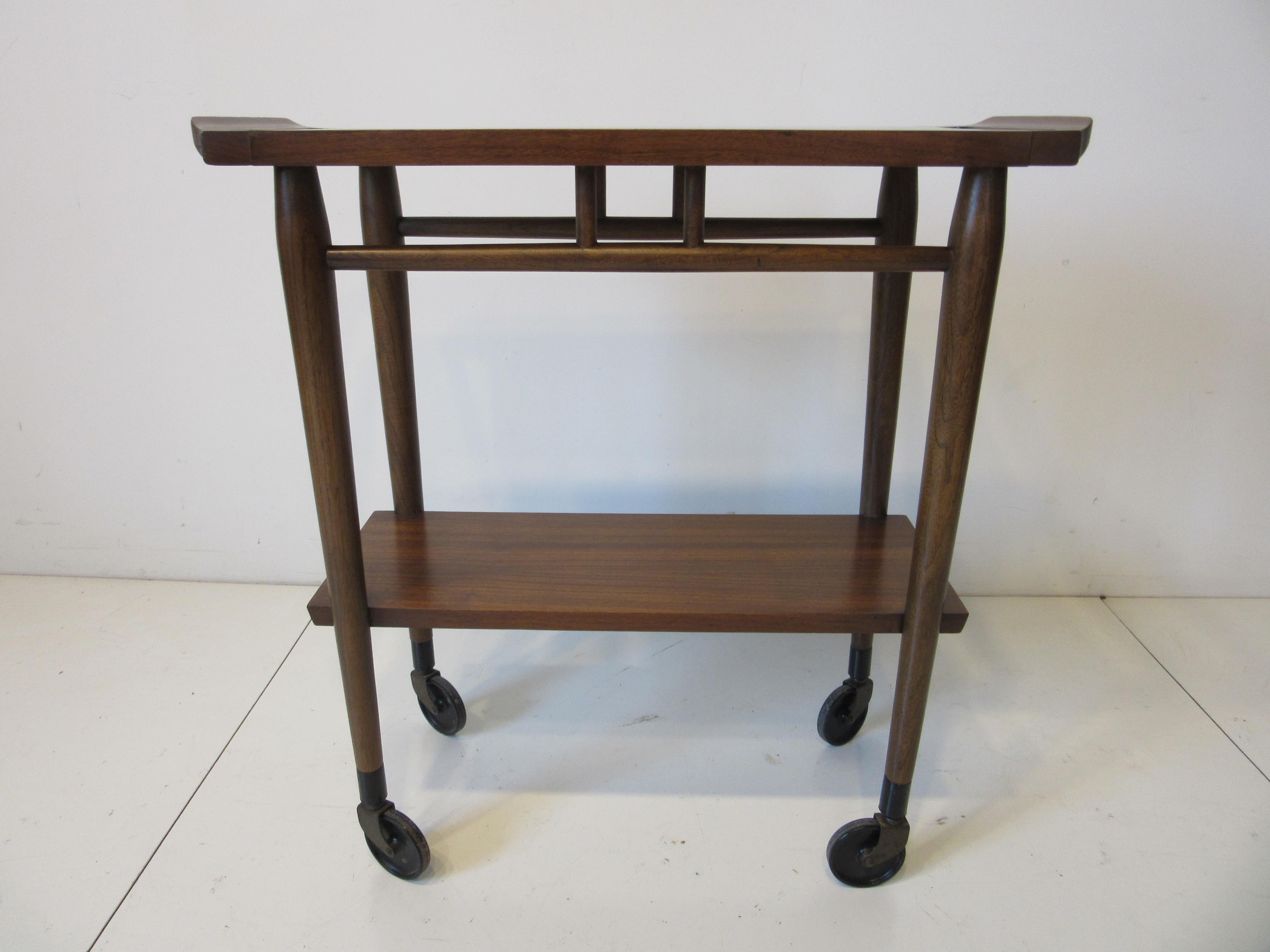 A nice smaller sized walnut rolling bar cart with cut out handles to the top and a lower shelve to the bottom sitting on conical black tipped legs with wheels. Branded mark to the verso of the shelve manufactured by the Lane Furniture Company. This