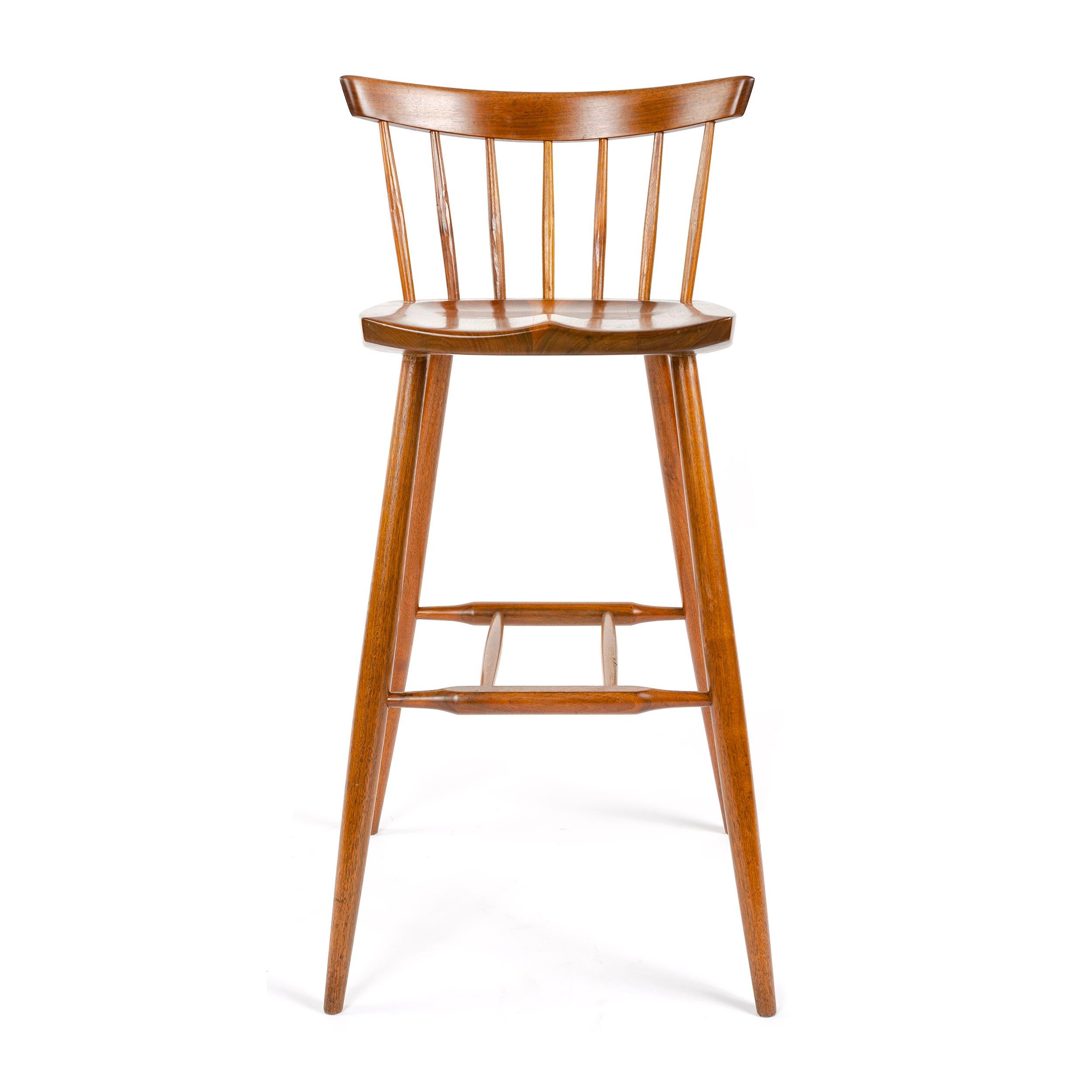 A walnut barstool designed by American Craftsman Master George Nakashima with hand-hewn spindles supporting a thin backrest, a shaped seat on four splayed, and tapered dowel legs with a footrest stretcher.
Made by the George Nakashima Studio in the