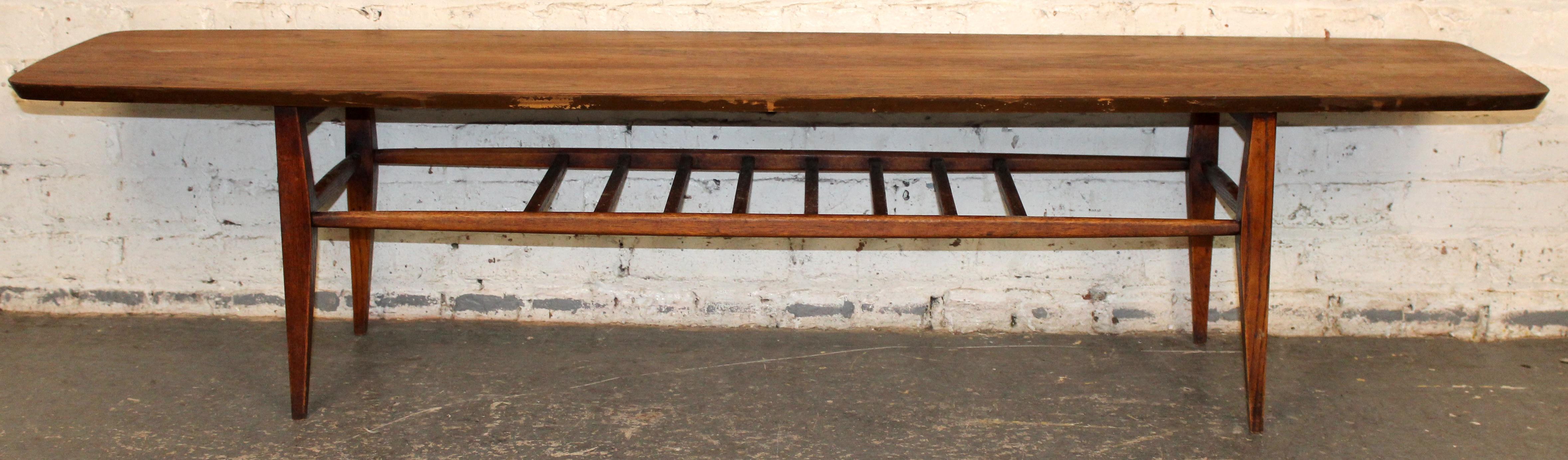 A classic mid-century surfboard-form coffee table crafted in warm walnut, featuring elegantly tapered ends and a long, sleek profile. Below the top rests a finely spaced spindled stretcher shelf, ideal for books or magazines while lending the piece