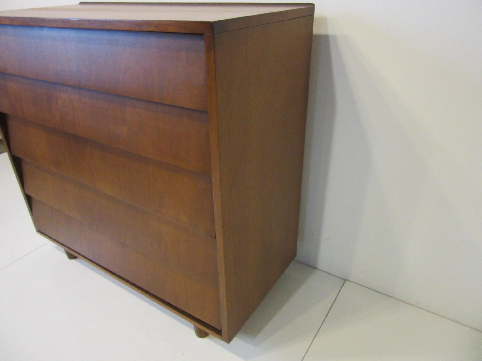 A walnut tall dresser / chest with five drawers with slanted fronts and a raised lip to the top back edge in the manner of Florence Knoll sitting on rounded legs, storage that makes a statement.