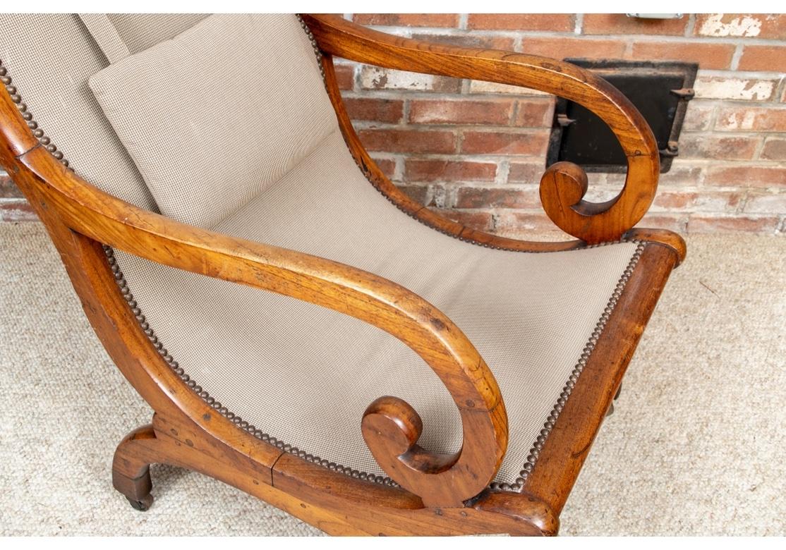 An Antique lolling chair with particularly attractive Amber Toned Walnut frame. With the classic sloping back, scrolled arms and curved legs. Upholstered in a brown and taupe tiny check fabric with attached lumbar cushion. Raised on casters. 
H. 38