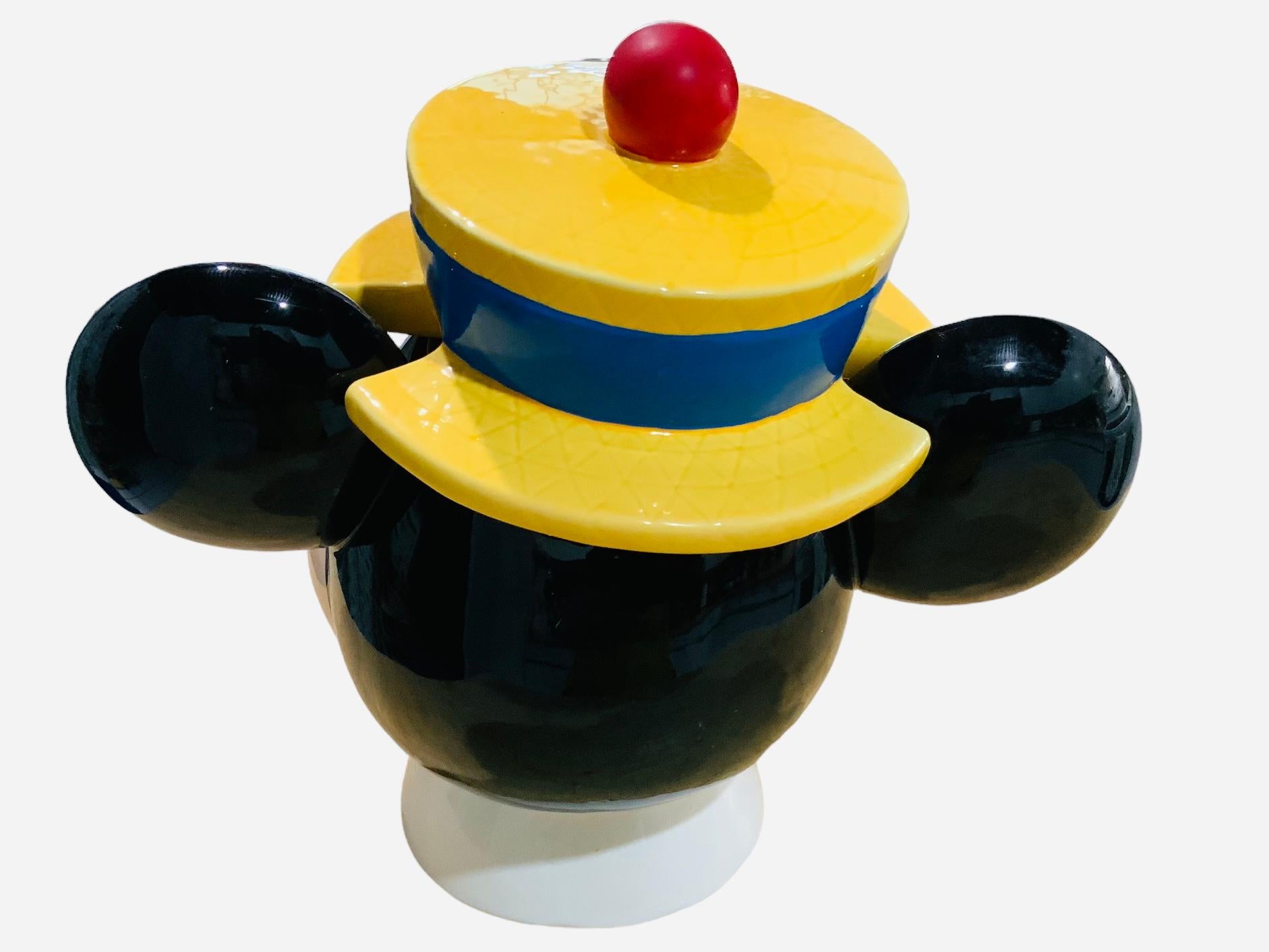This is a Walt Disney, Mickey Mouse Head Cookie Jar. It depicts a hand painted glazed ceramic cookie jar shaped as Mickey Mouse head. He is wearing a yellow hat with a band that have written Cookies. Below the base is hallmarked C Disney.