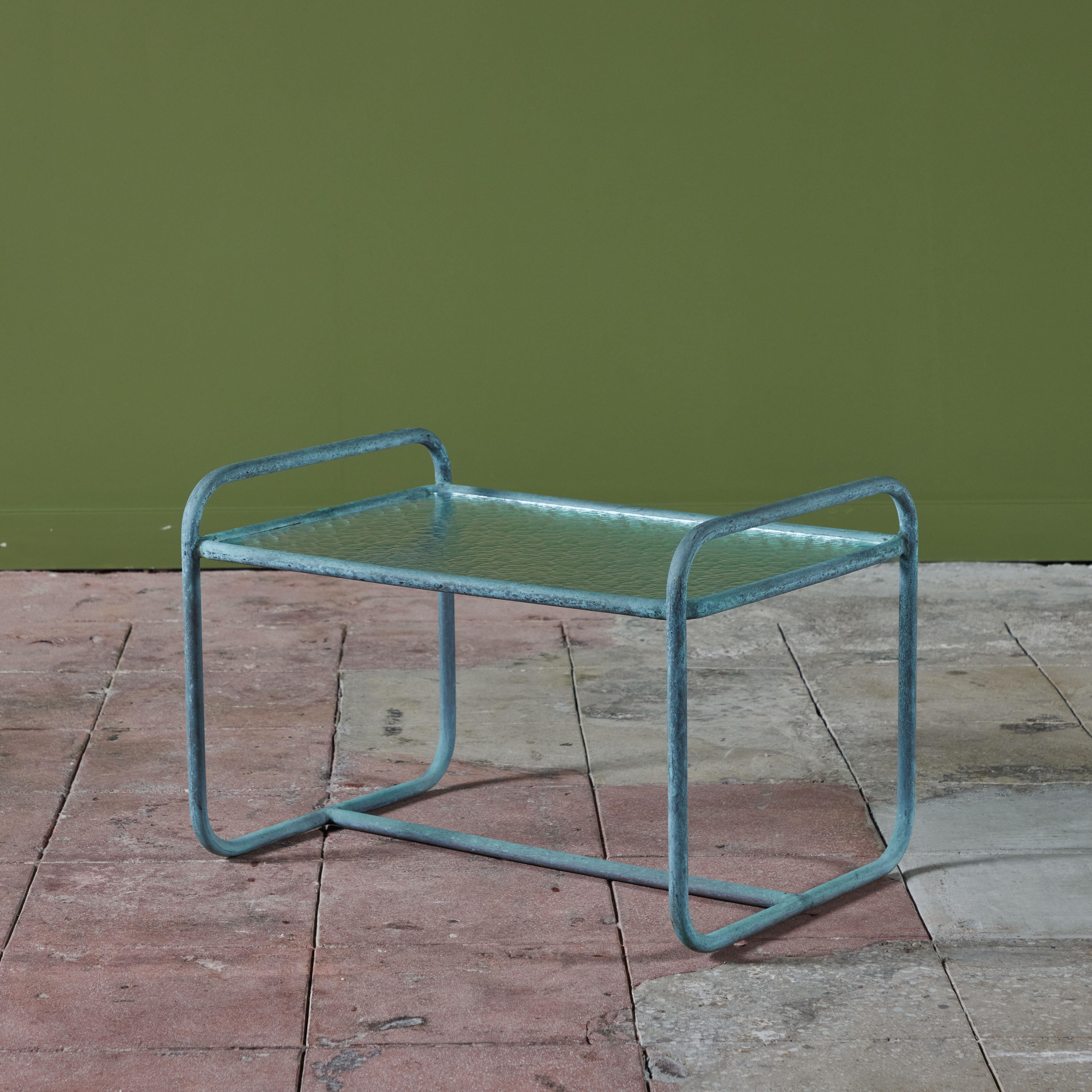 Patio side table designed by Walter Lamb and produced by Brown Jordan. The table frame is bronze tubing with a verdigris patina with an inset hammered glass tabletop. Two curved rails form the legs and create the “handles” on either side of the