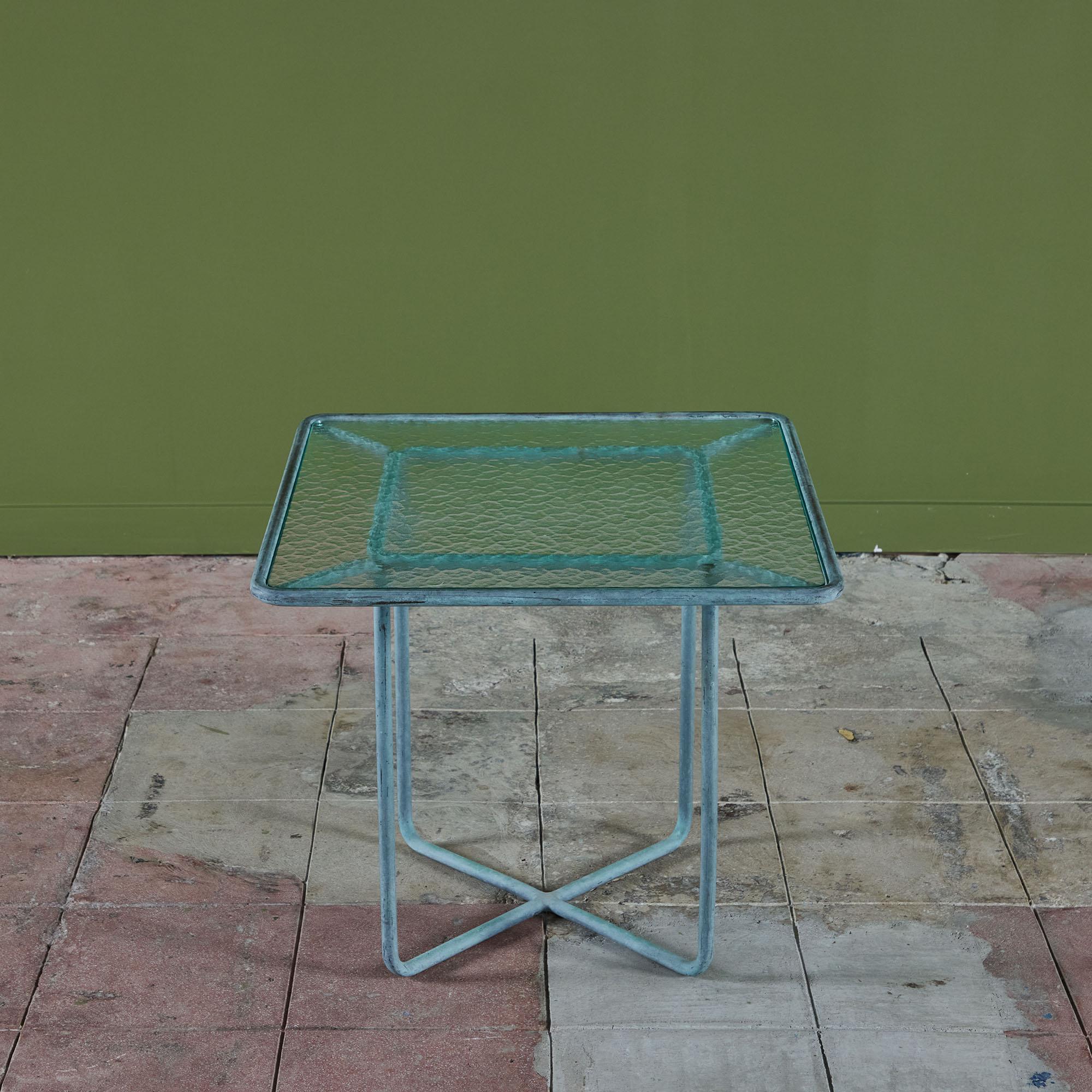 A side table in patinated bronze designed by Walter Lamb and produced by Brown Jordan. The table has a square shape with rounded corners, supported by four legs in matching bronze. The legs intersect with horizontal bars that sit on the ground for