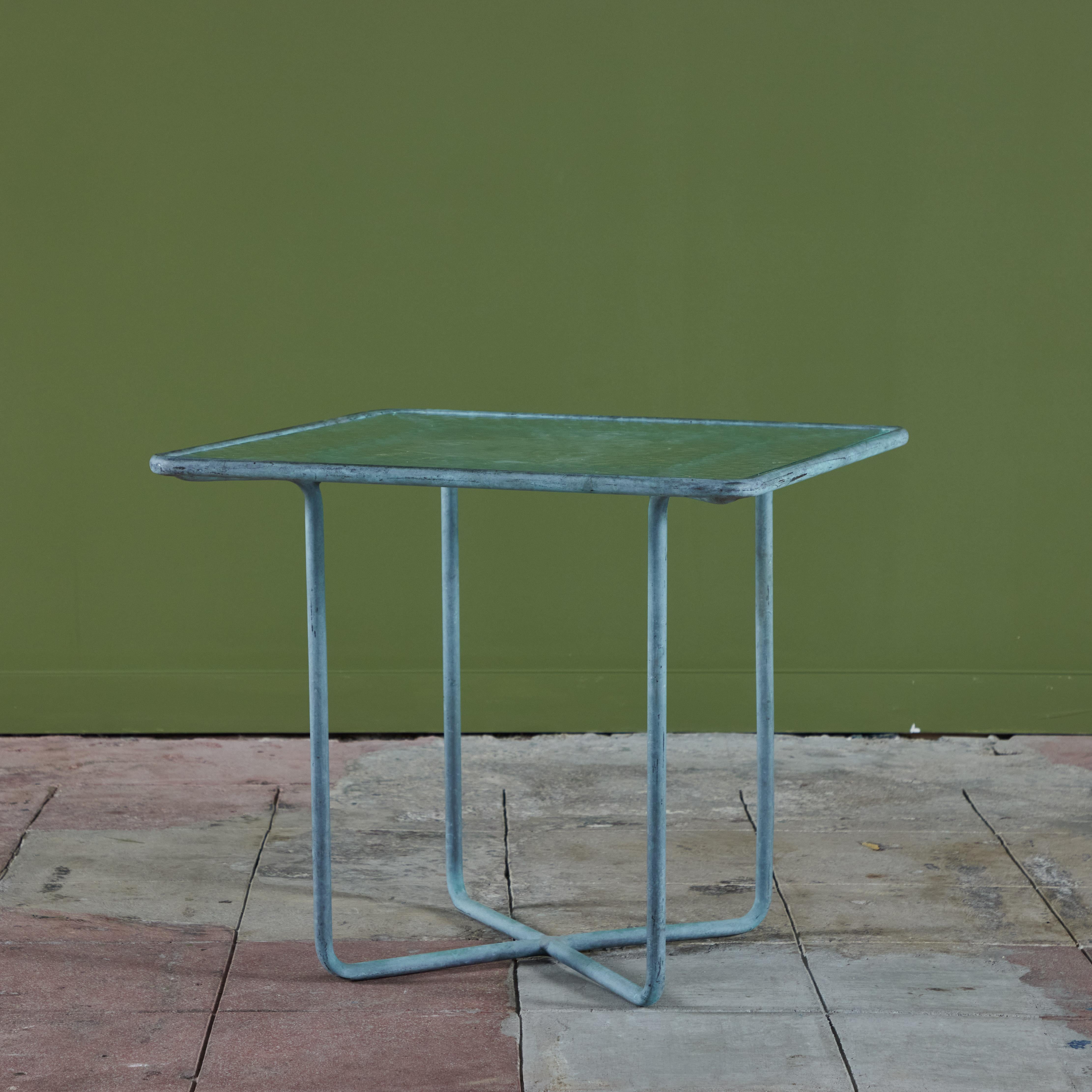 Mesa auxiliar de bronce patinado diseñada por Walter Lamb y producida por Brown Jordan. La mesa tiene forma cuadrada con esquinas redondeadas, y se apoya en cuatro patas de bronce a juego. Las patas se cruzan con barras horizontales que se apoyan en