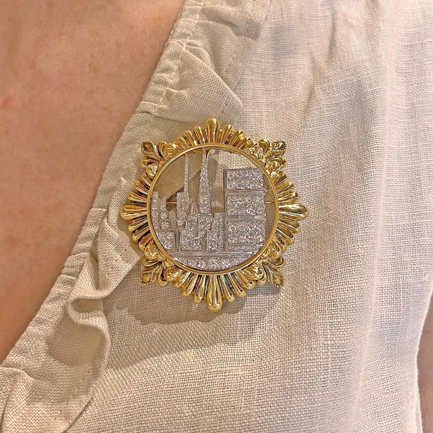 Pendant brooch, centering round-cut and baguette-cut diamonds set to resemble a city skyline in platinum surrounded by an 18k yellow gold fluted frame with four fleur-de-lis motifs.  Reverse has a curved hook that allows the wearer to suspend it on