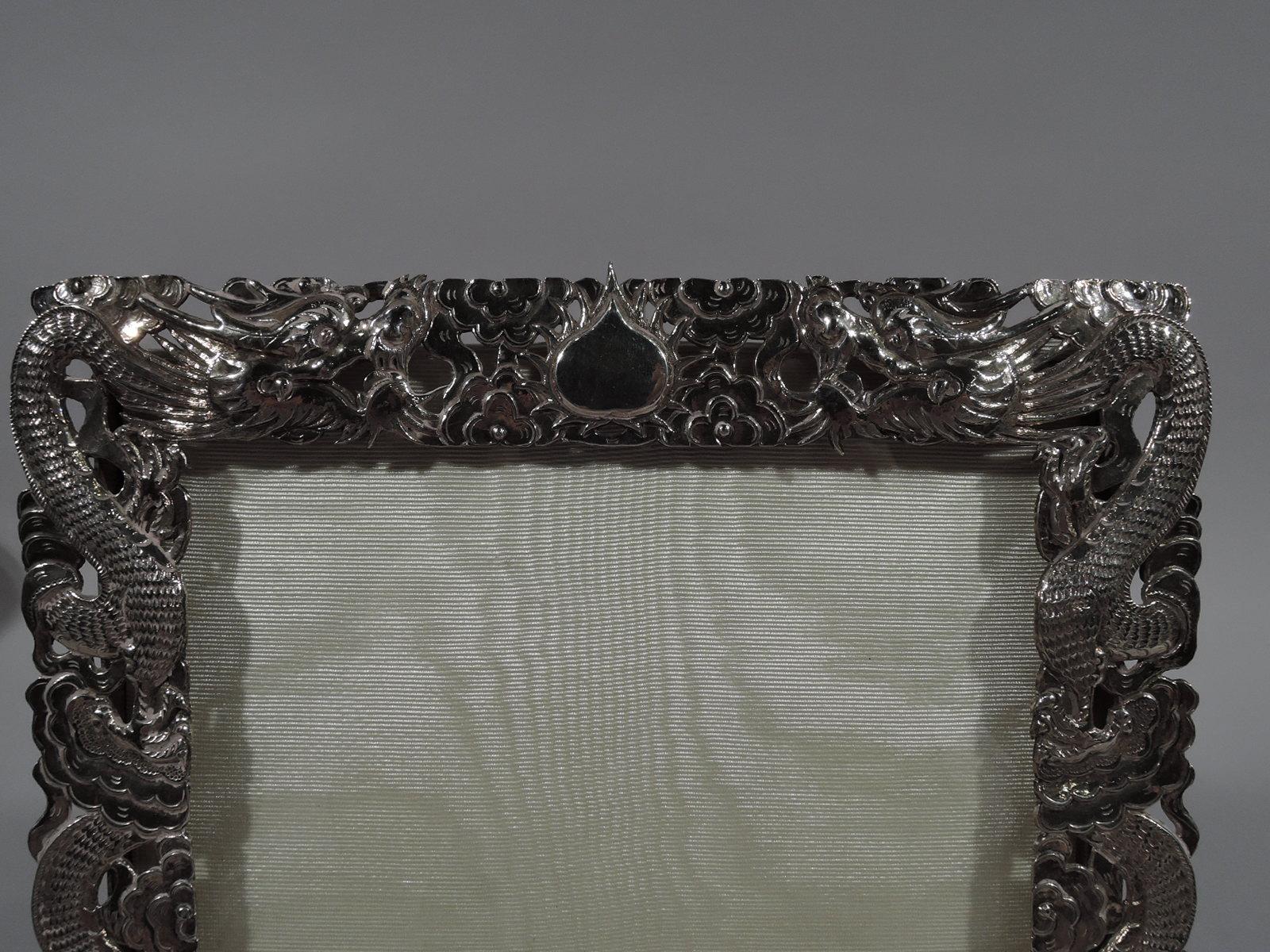 Chinese export silver picture frame, circa 1910. Rectangular window in pierced surround made up of serpentine serpent-dragons slithering through clouds. At top mono plate (vacant). With glass, silk lining, and bamboo-style wire hinged support.