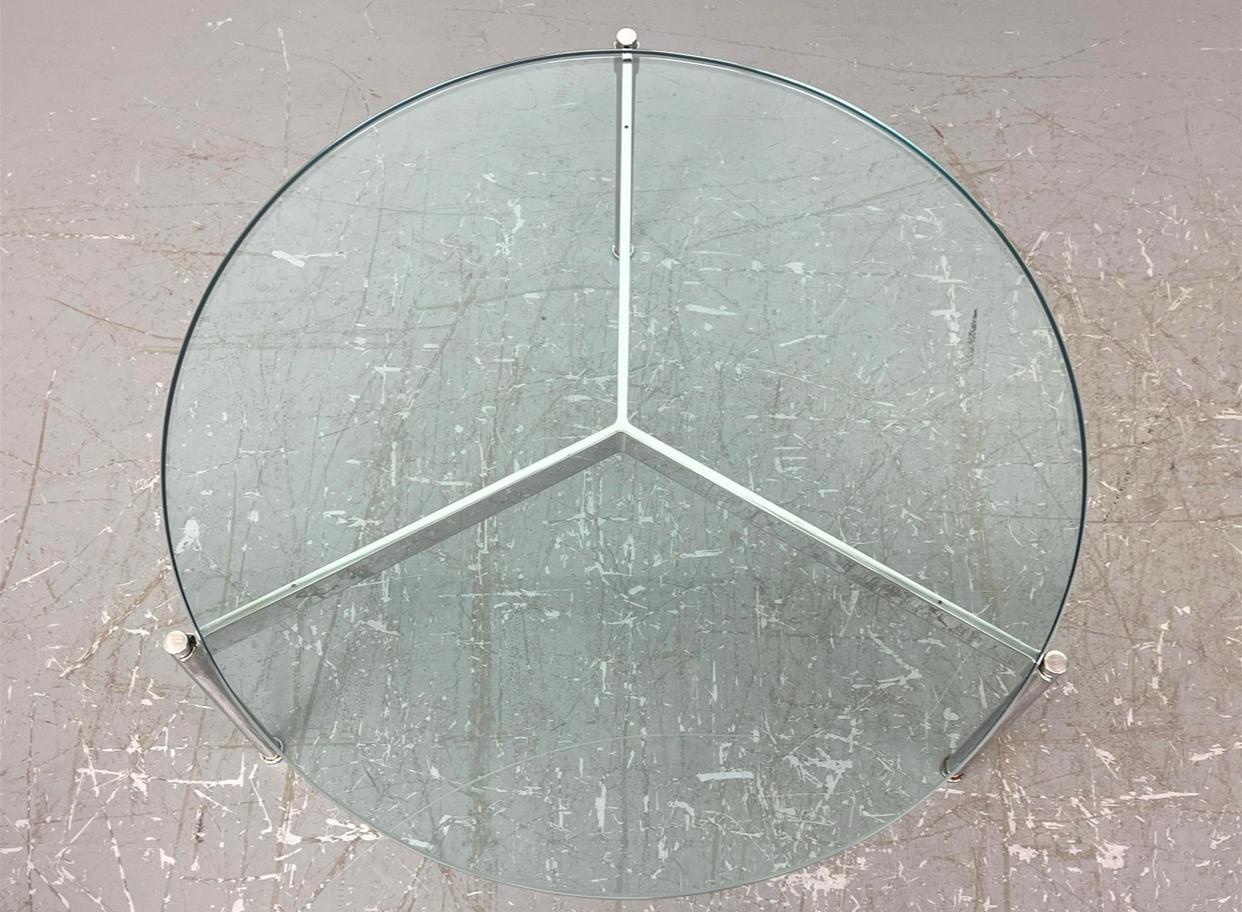 Circular cocktail table comprised of a glass top supported by a polished steel three legged base. The glass sits on a triform steel stretcher with claw like ends where it meets the legs. That is how the table, designed by Ward Bennett and