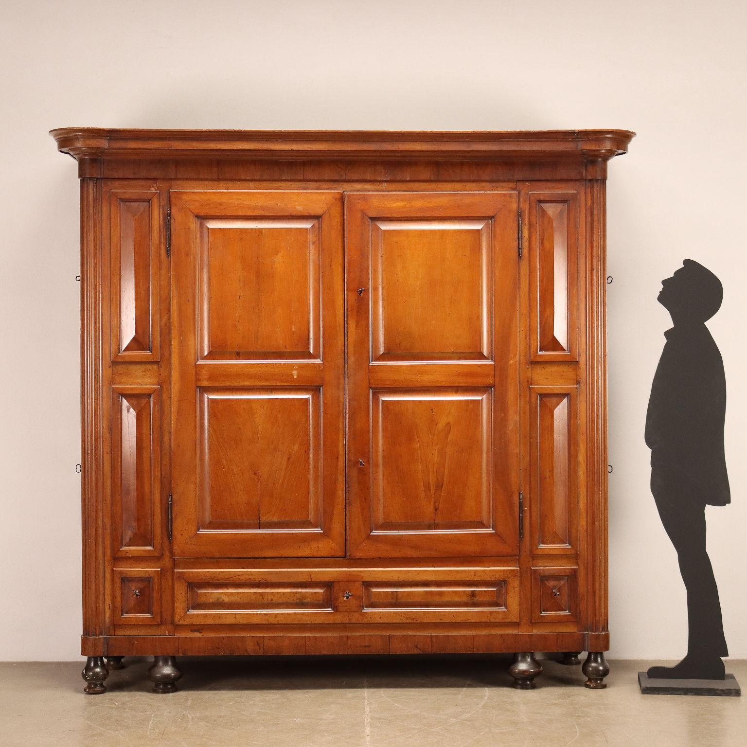 Empire wardrobe in walnut, Italy, first quarter of the 19th century. Molded frieze, front with a pair of doors and lower band with 3 drawers, tapered columns on the uprights, sides house 4 pantry compartments, turned feet. Base and top are connected