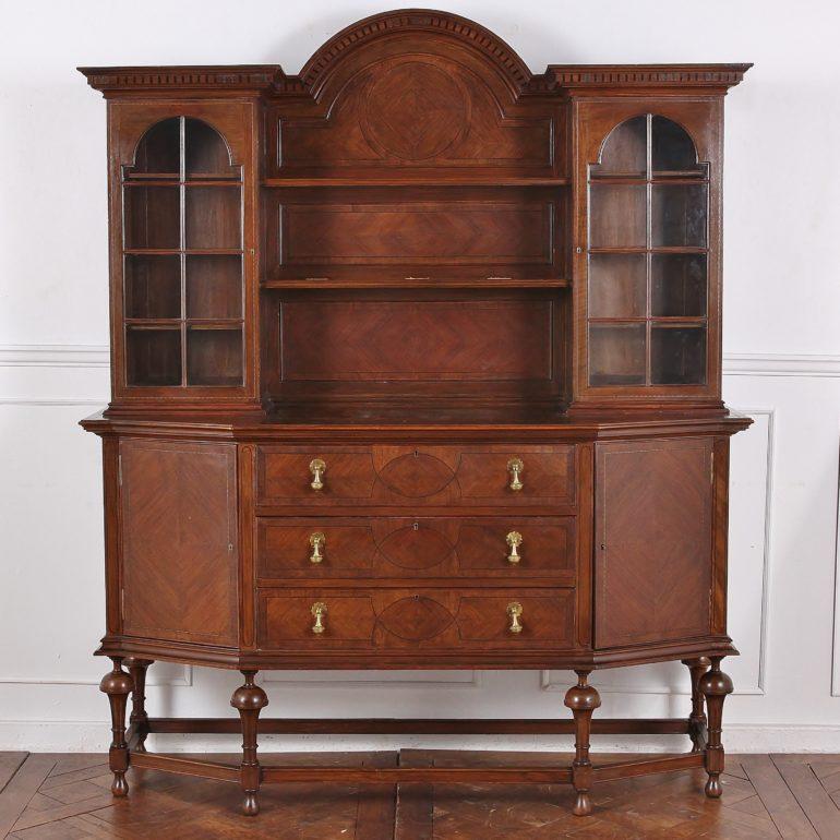 'Waring & Gillows' English buffet, the base with angled side cabinets flanking a bank of three drawers and raised up on barrel-turned legs united by a stretcher. The upper cabinet offers central open shelves between two side glass-fronted arched