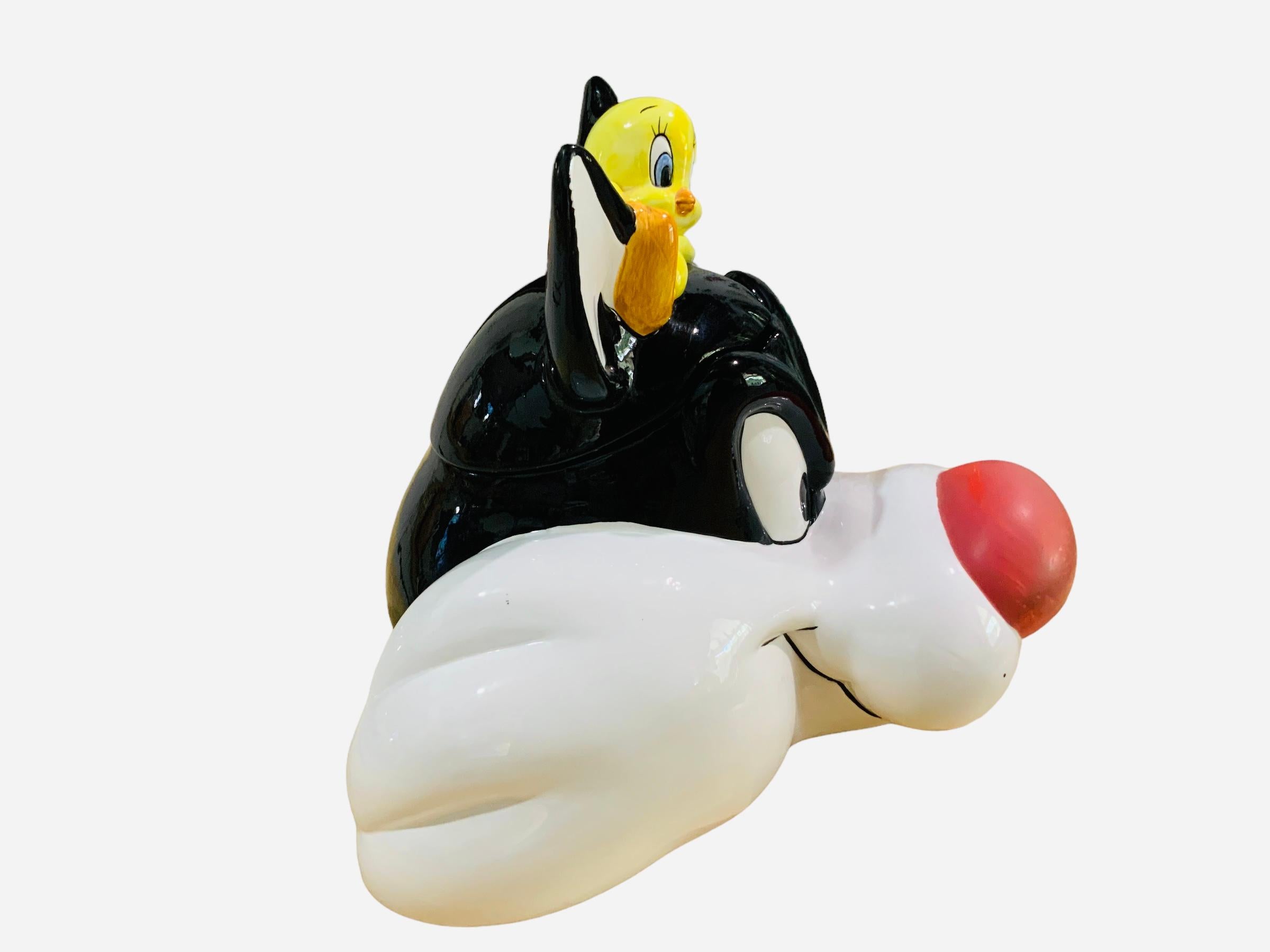 This is a Sylvester and Tweety Bird Cookie Jar. It depicts a hand painted glazed ceramic cookie jar shaped as the head of Sylvester and Tweety Bird laying down above it. Below the base is hallmarked Applause, Inc.; Warner Bros. Looney Tunes.