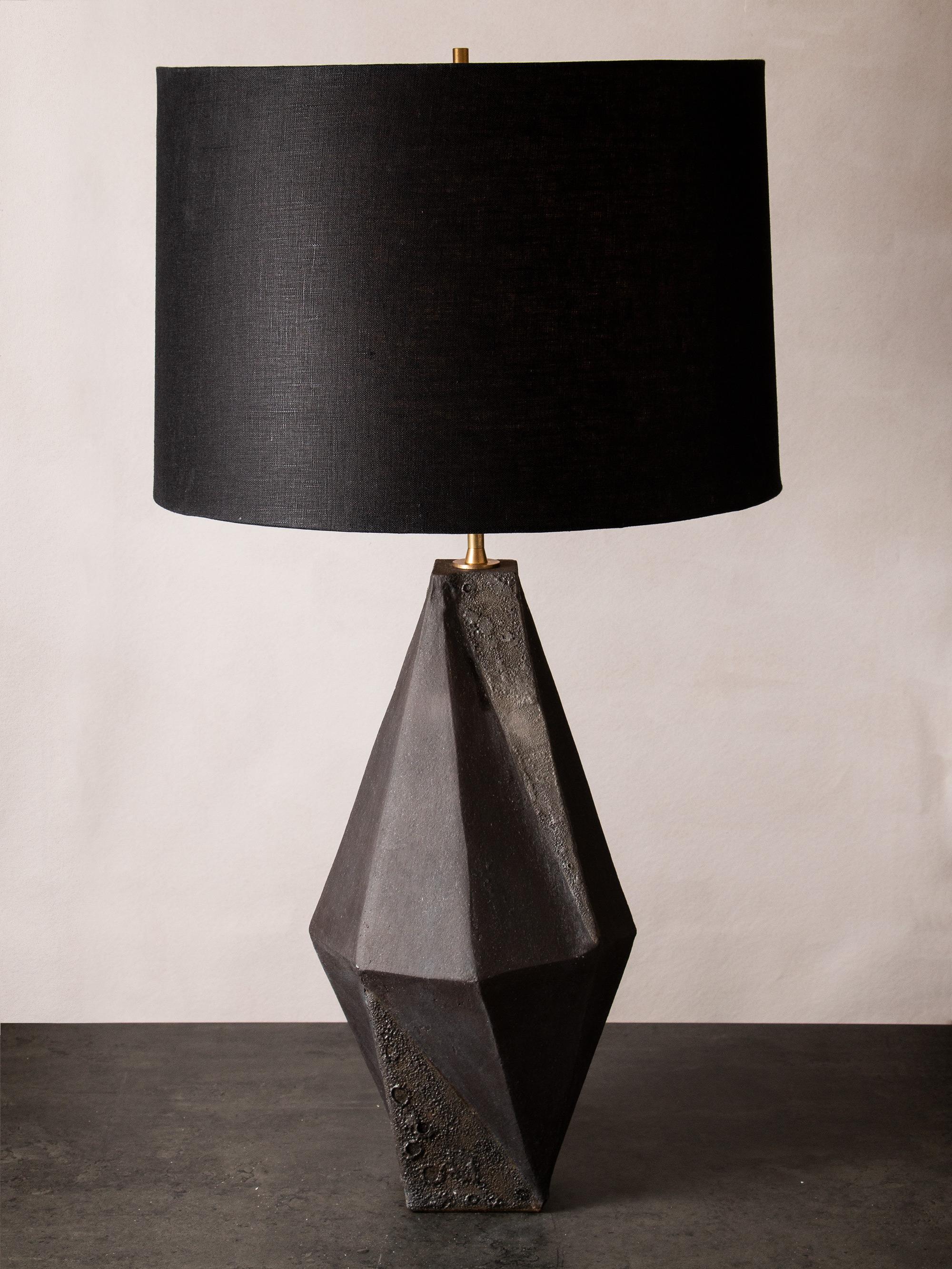This dramatic black ceramic table lamp features a complex geometric shape, accentuated by contrasting matte black and textured black glazes. Each piece is individually handmade and entirely unique. The lamp is finished with raw brass hardware and a