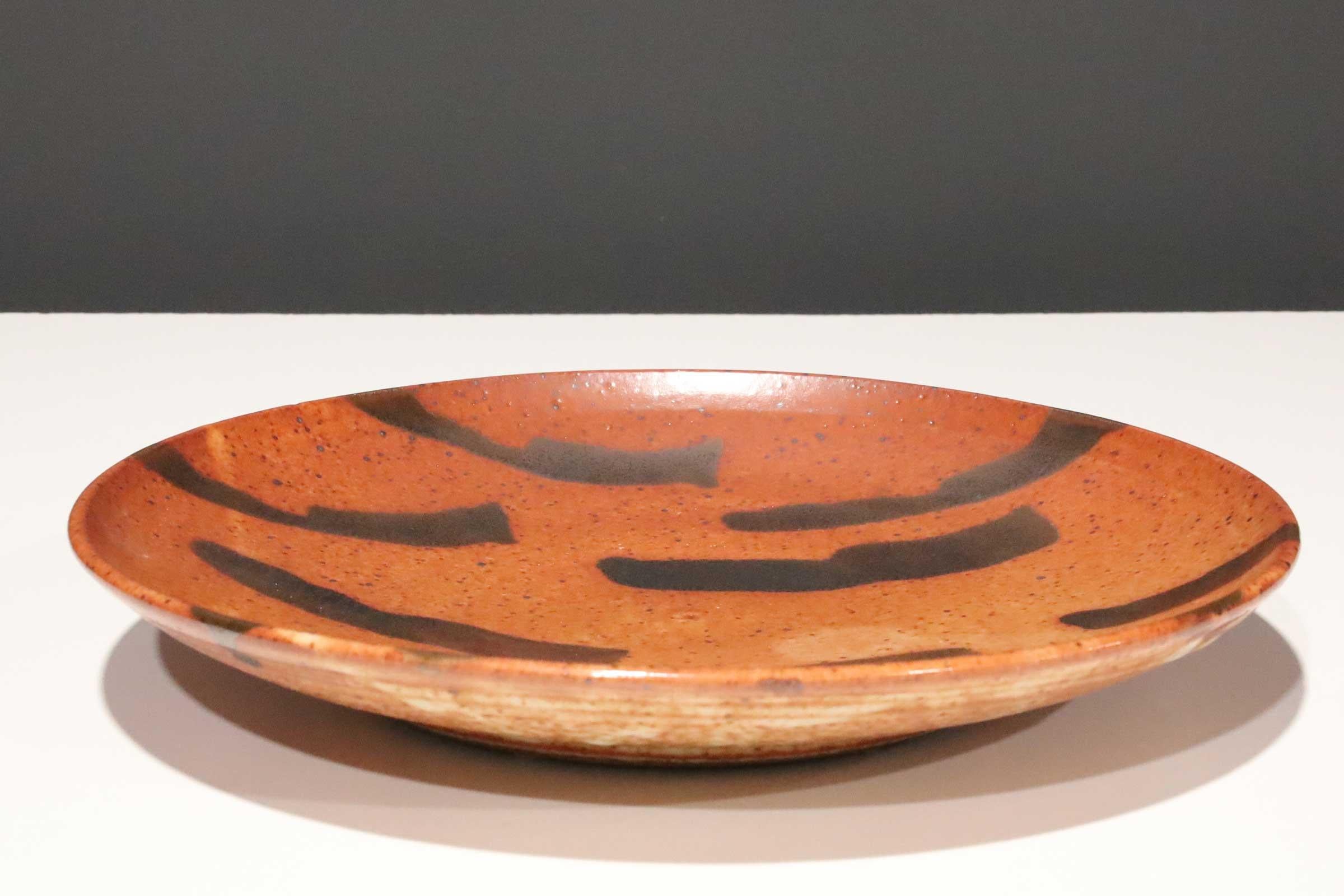 Large studio pottery platter with a red glaze and brown decorative dashes. 

Warren MacKenzie is known for simple, wheel-thrown functional pottery influenced by Bernard Leach and the Japanese aesthetic of the work of Shoji Hamada.

In 1950