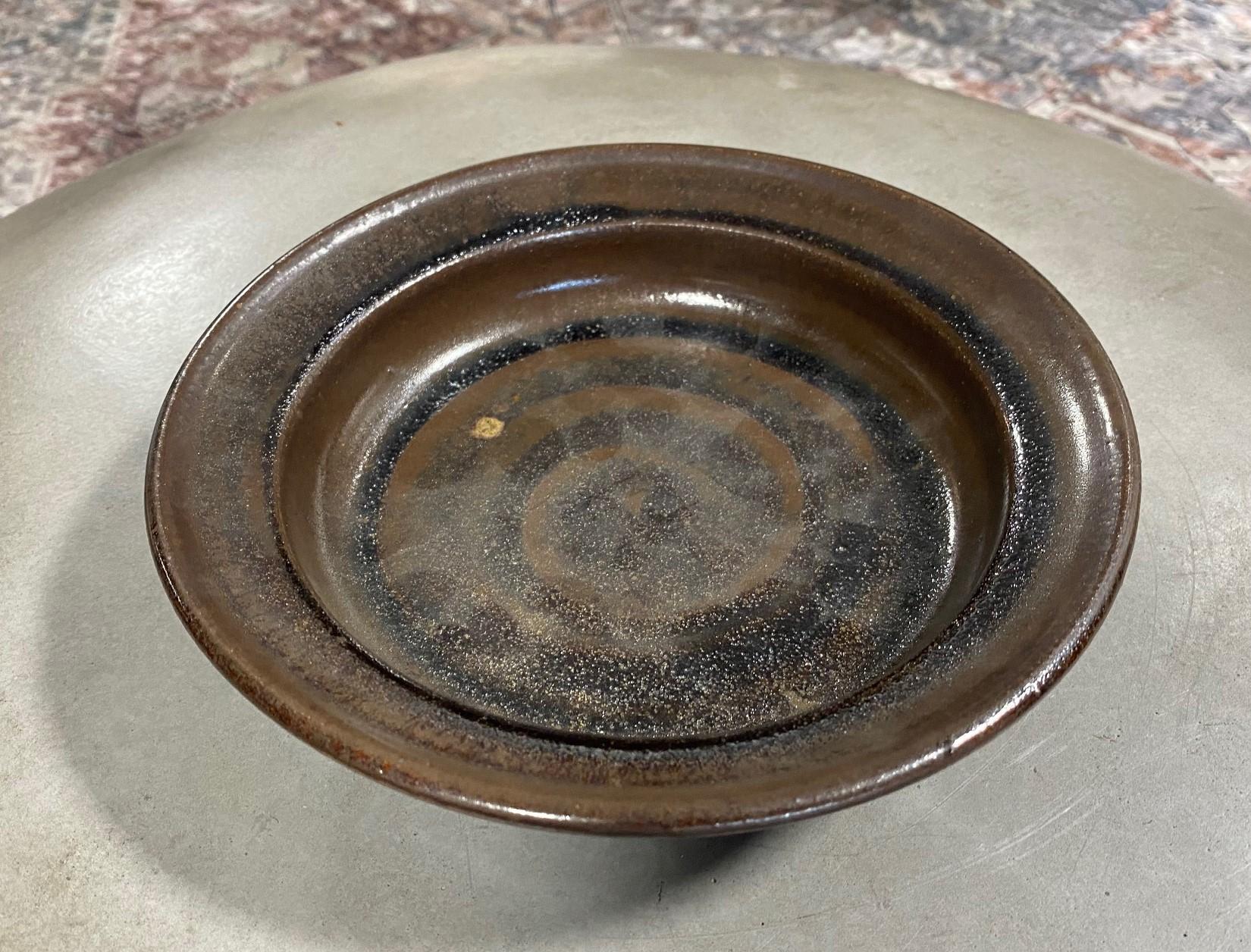 A gorgeous work by 20th century master American and renowned Minnesota studio potter/artist Warren MacKenzie. 

The three-footed tenmoku glazed bowl is signed/ stamped along the foot rim by MacKenzie and features a unique decorative pattern. The