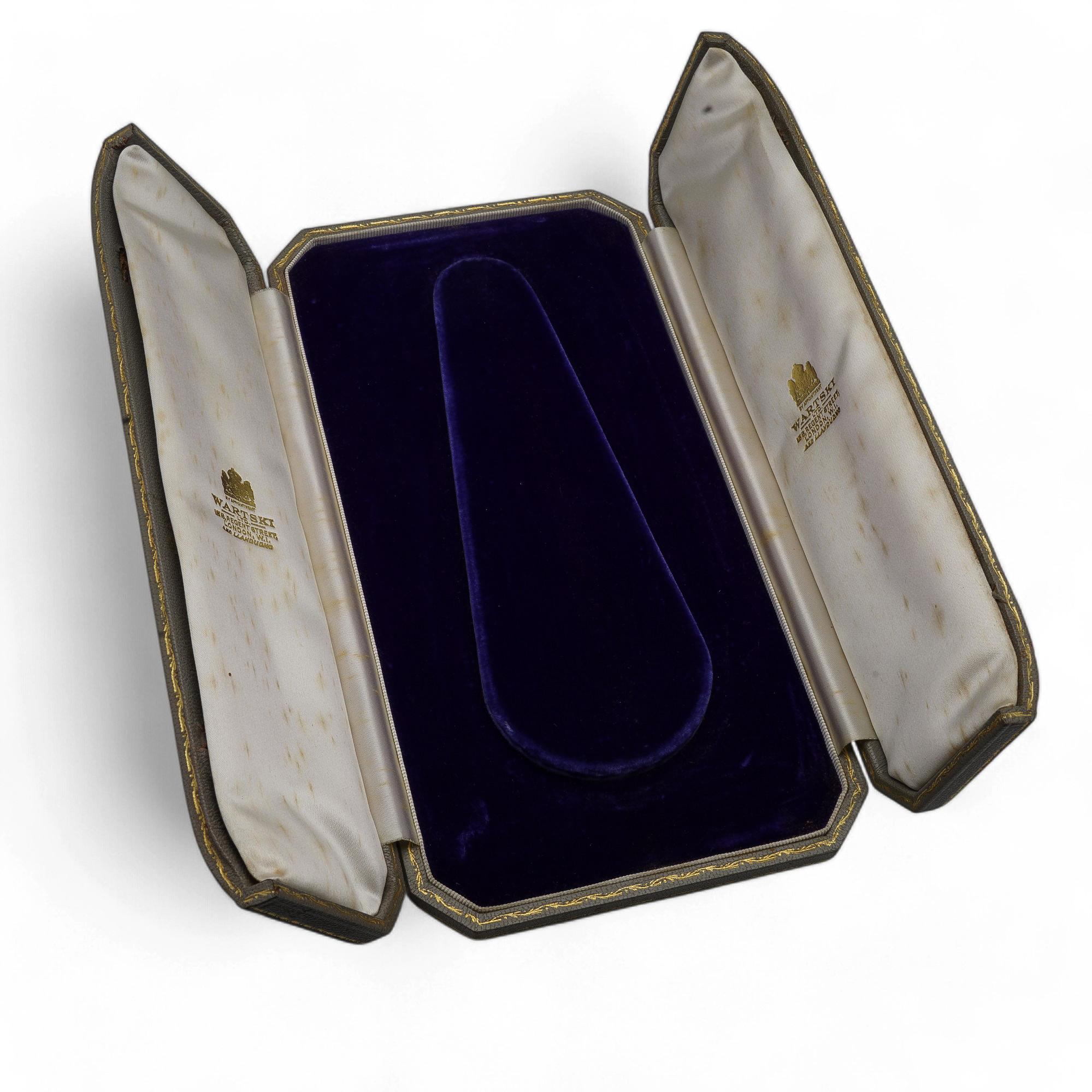 Une boîte à collier rectangulaire avec un extérieur texturé et un intérieur en velours conçu pour accueillir un collier en toute sécurité. Les clips sont manquants, ce qui affecte la fermeture.

Pièce ancienne de Wartski, cette boîte à colliers
