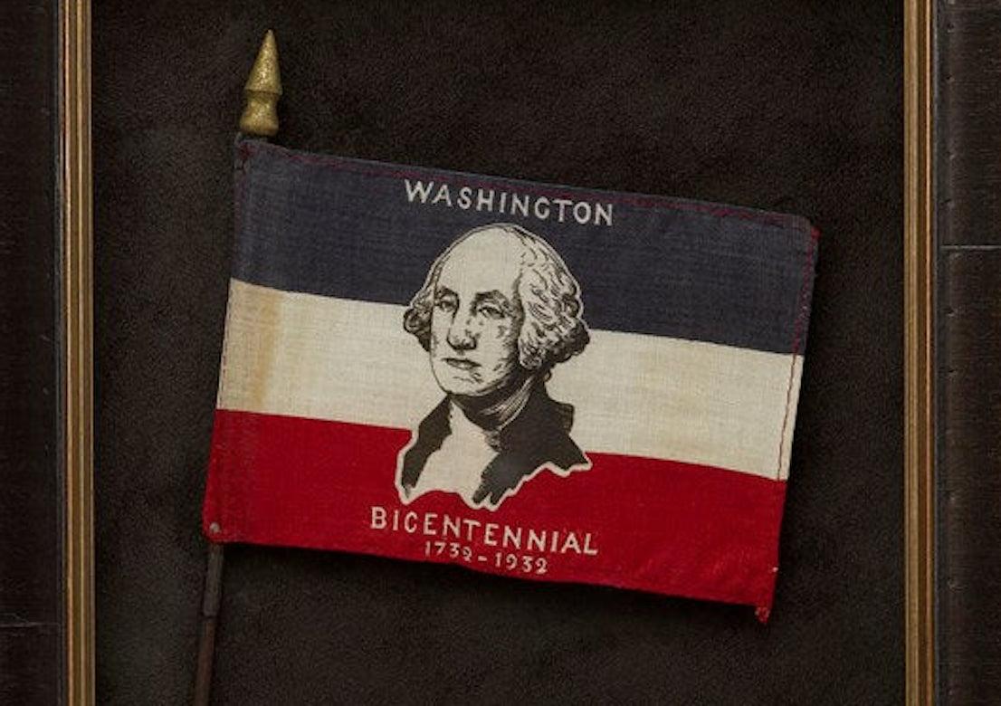 Presented is a George Washington Bicentennial parade flag from 1932. The flag is printed with blue, white, and red stripes, with a prominent bust portrait of George Washington at center. The words “Washington Bicentennial” and the dates “1732-1932