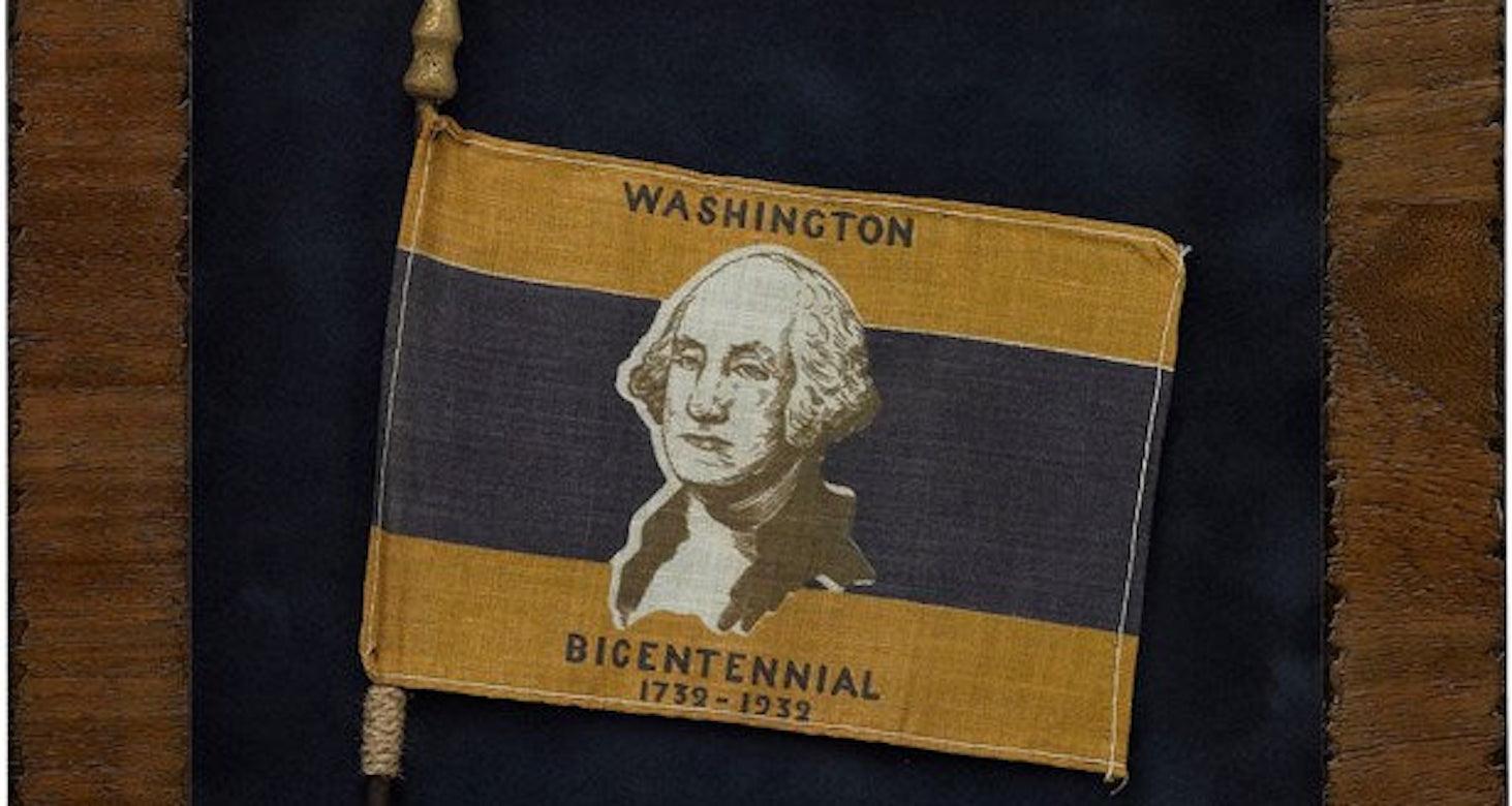 Presented is a George Washington Bicentennial parade flag from 1932. The flag is printed with gold, white, and blue stripes, with a prominent bust portrait of George Washington at center. The words “Washington Bicentennial” and the dates “1732-1932
