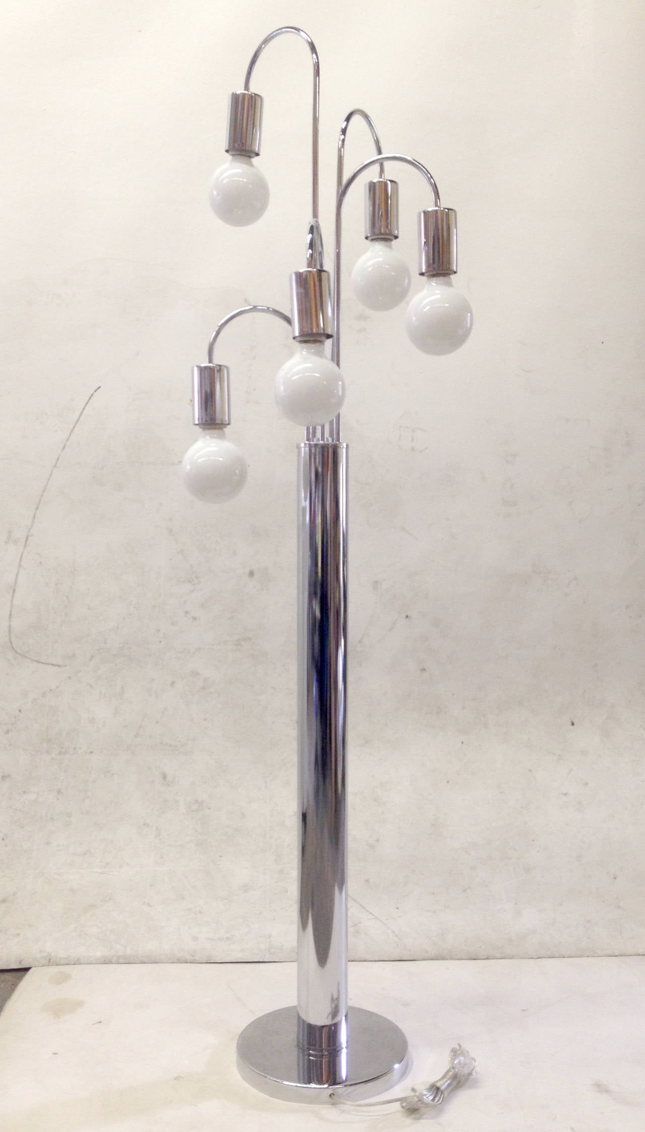This five-arm floor lamp has a very 1970s design rendered entirely in chrome. Five curved branches radiate from the central cylinder in a helix and each ends in an opaque white globe bulb. The lamp is operated by a dimming dial that controls the